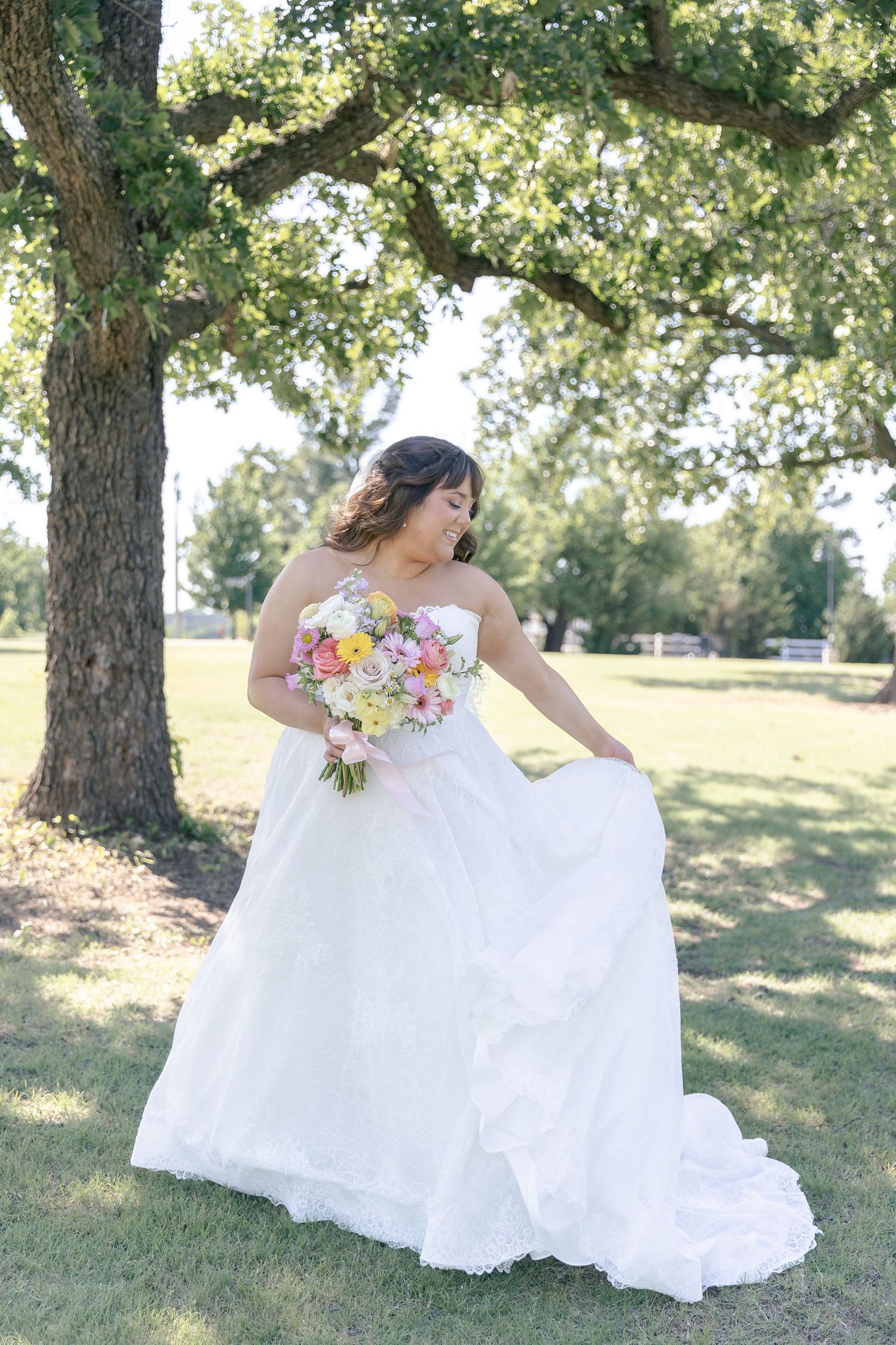 Bride in strapless white ball gown holding colorful bouquet, posing under oak tree on sunny day.