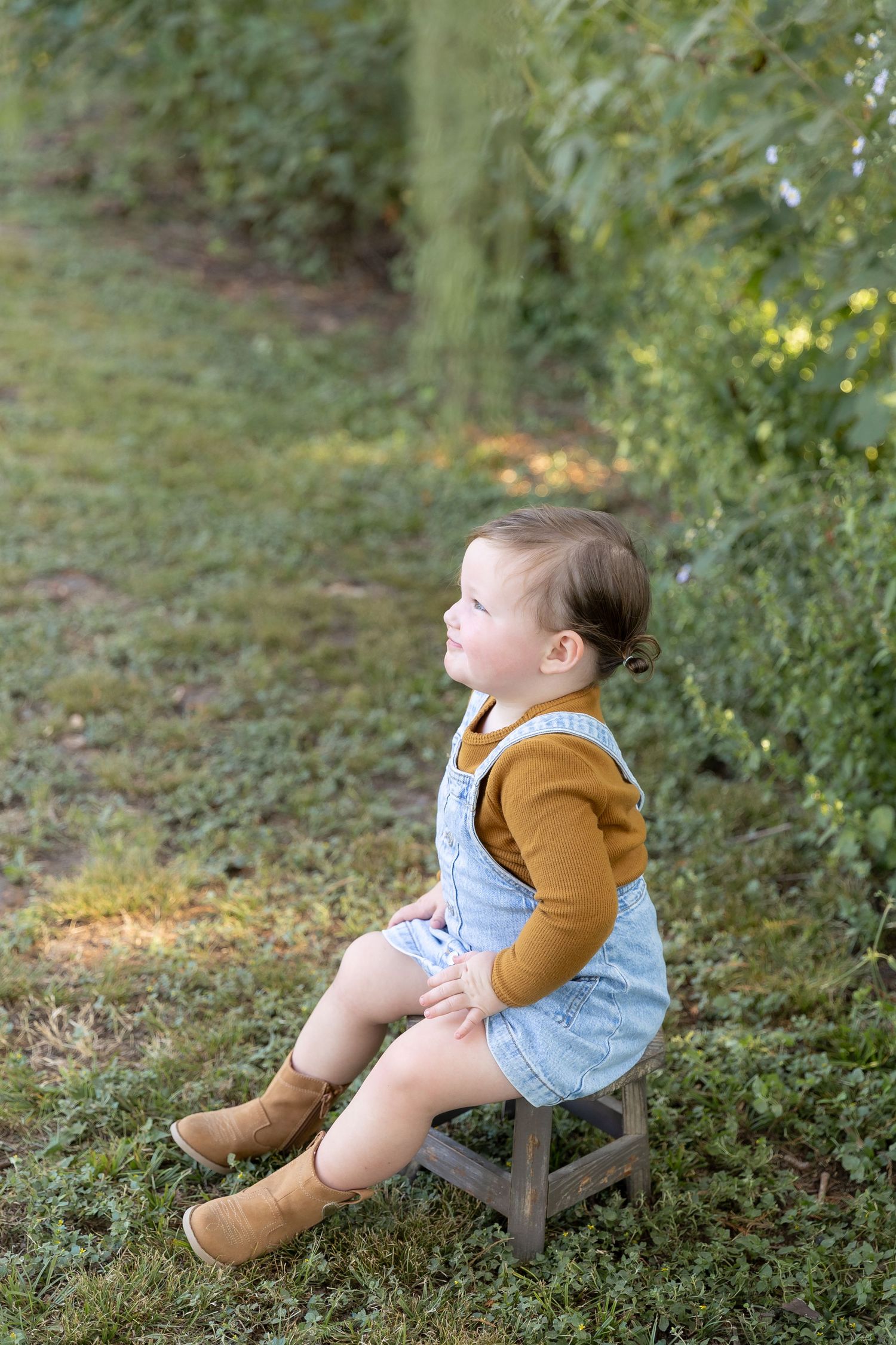Young child in overalls and brown sweater sitting on ground during outdoor photo session.