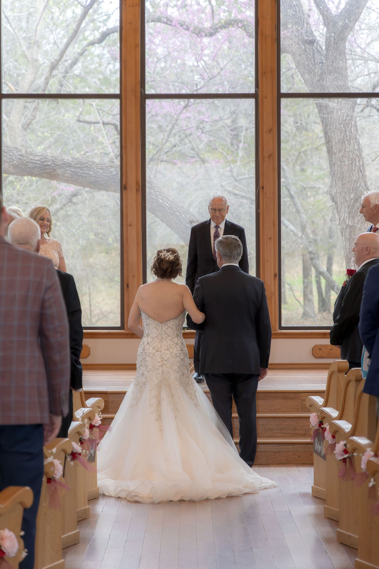 Bride and groom walk down the aisle in a chapel with large windows overlooking trees during wedding ceremony.