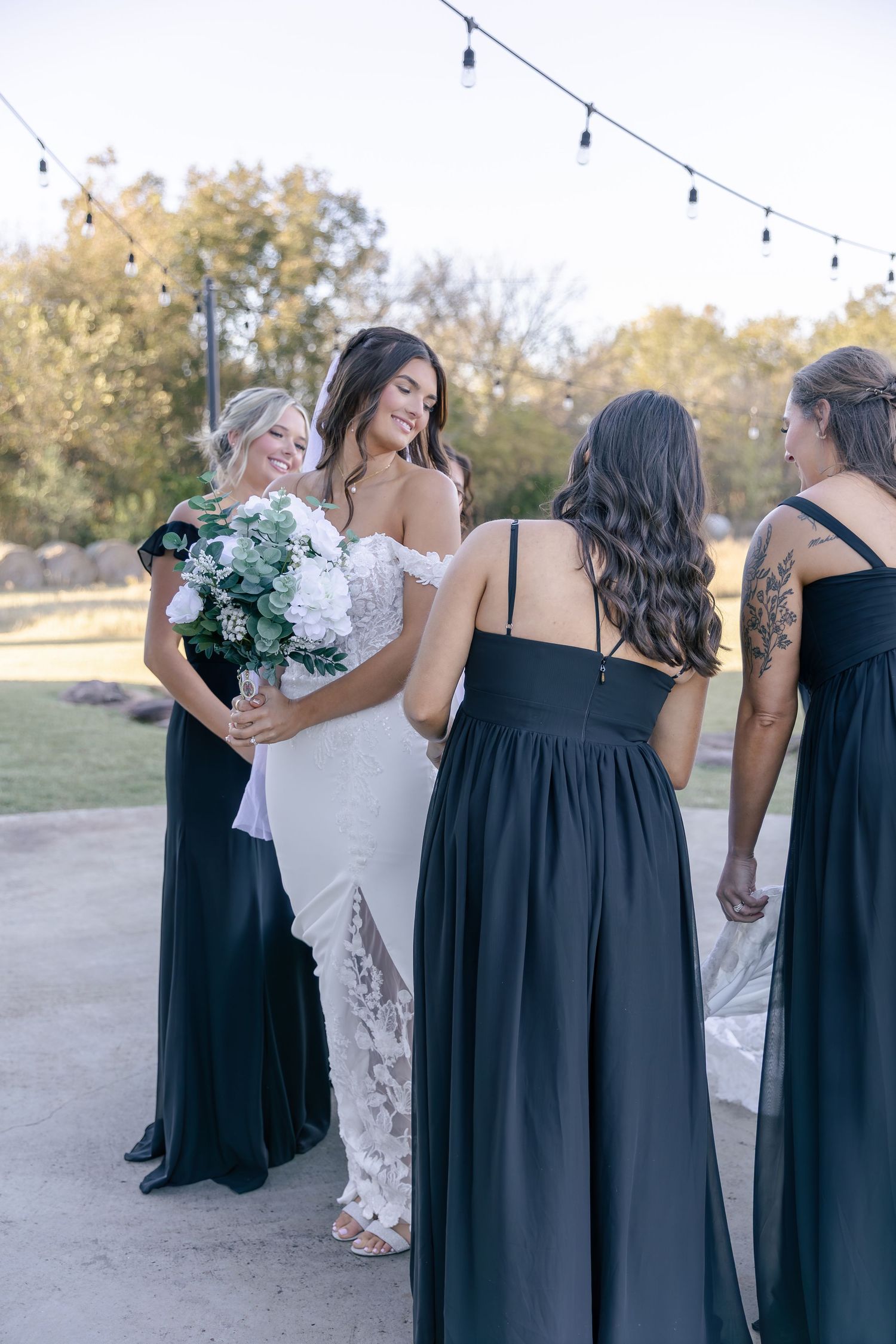 A wedding party in black bridesmaid dresses and a white bridal gown gathers outdoors under string lights at sunset.