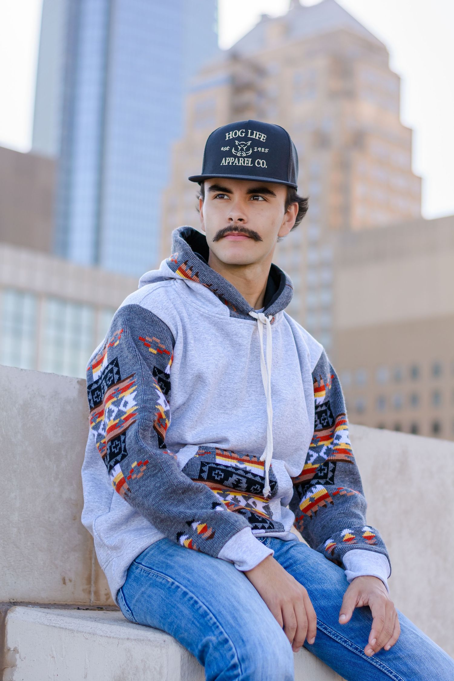 Casual urban senior portrait of a man wearing a baseball cap and a patterned jacket against a concrete background.