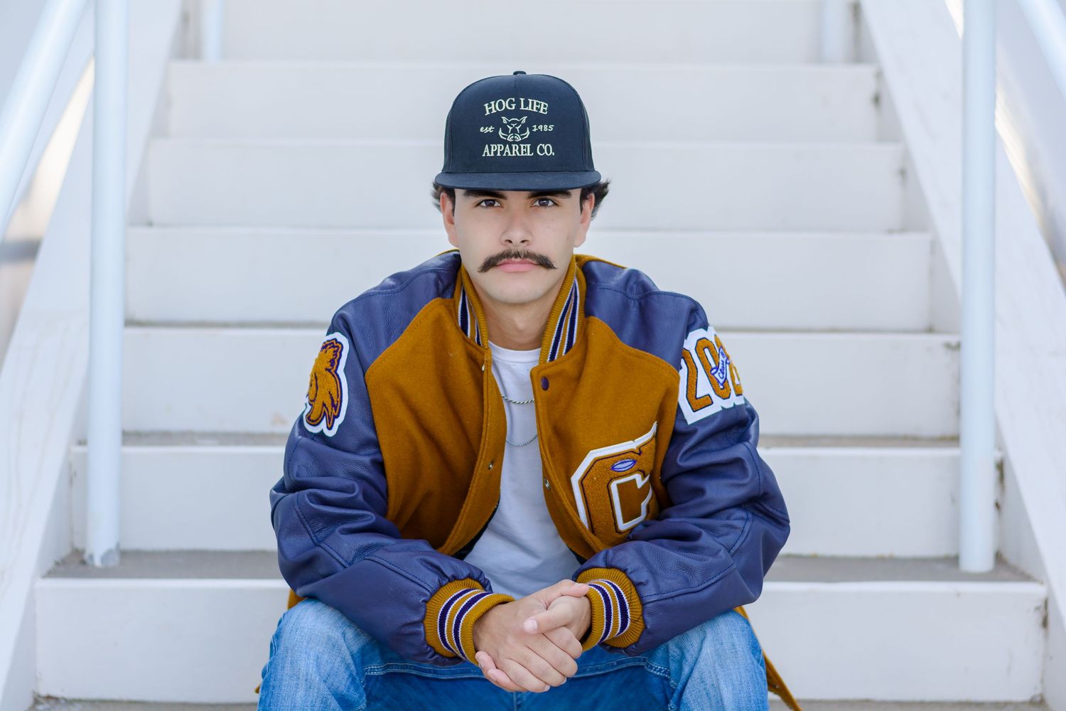 a senior in a varsity jacket and baseball cap sitting on white stairs.