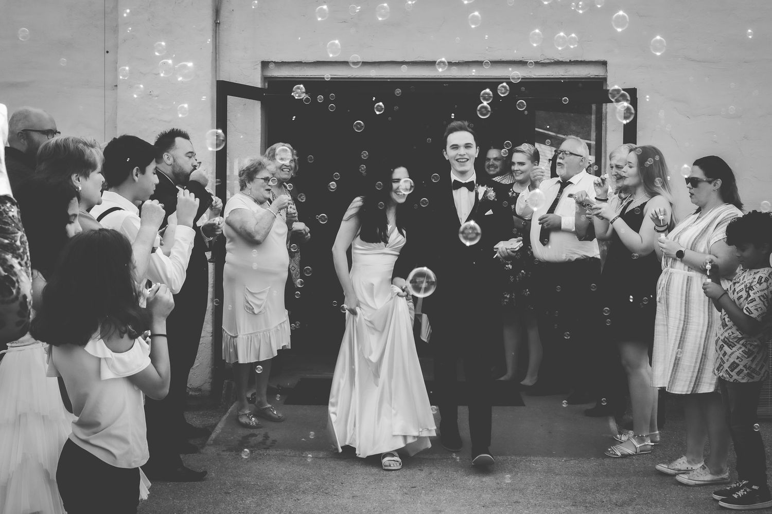 Wedding guests blow bubbles while celebrating during an outdoor reception in black and white.