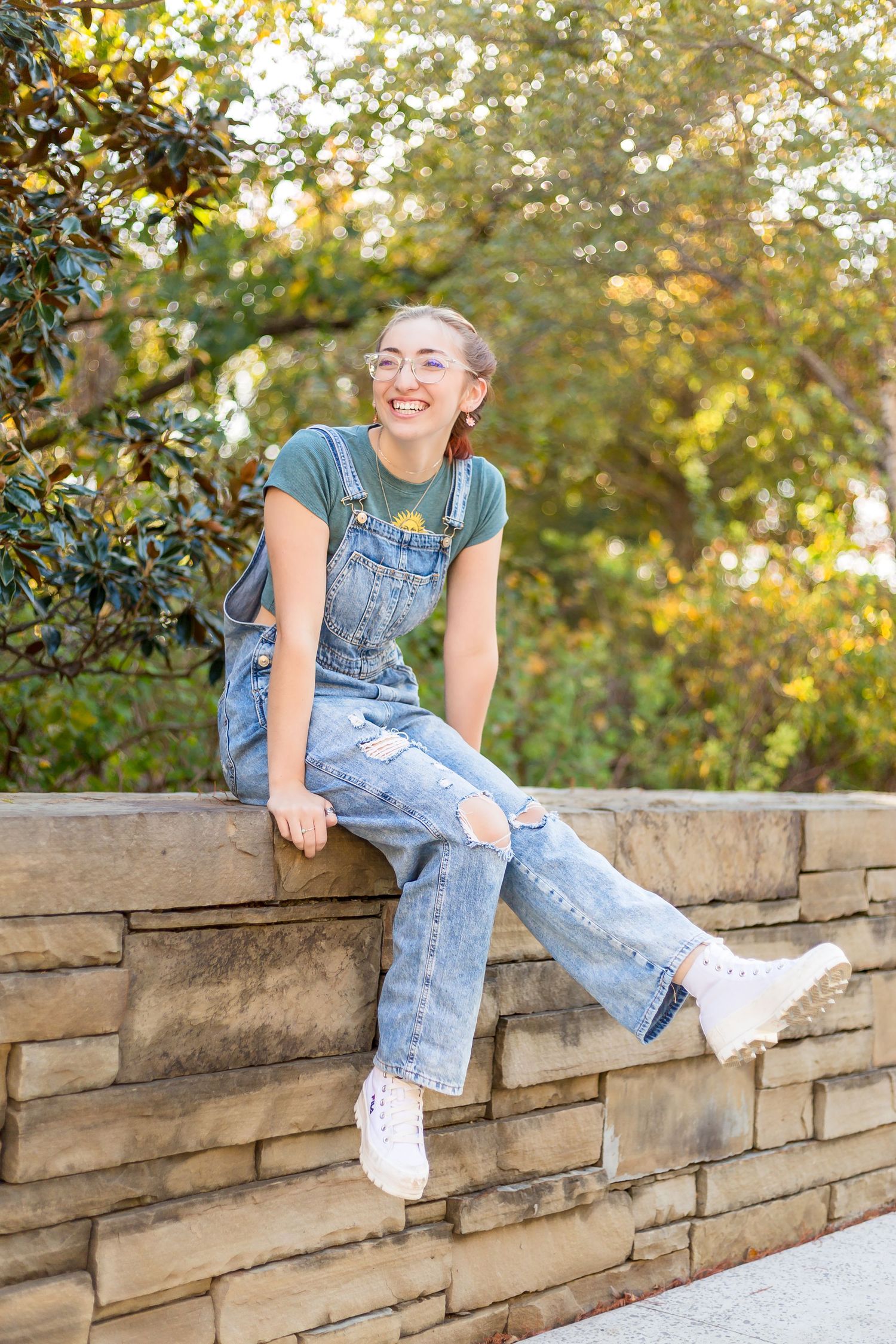 Candid outdoor senior photoshoot on a stone wall, wearing overalls and white sneakers at sunset.