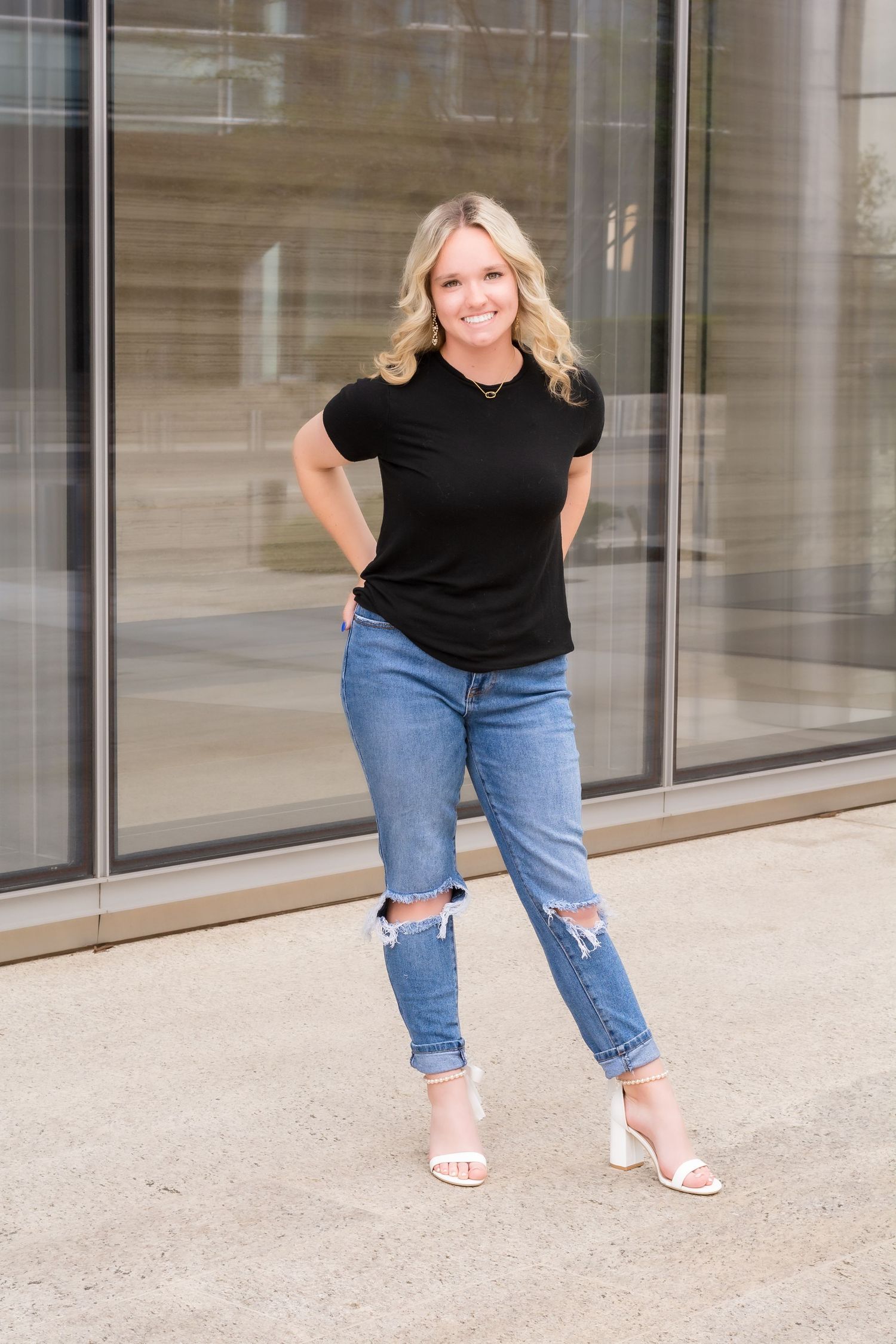 Casual senior poses in a black top and jeans outside a modern building.