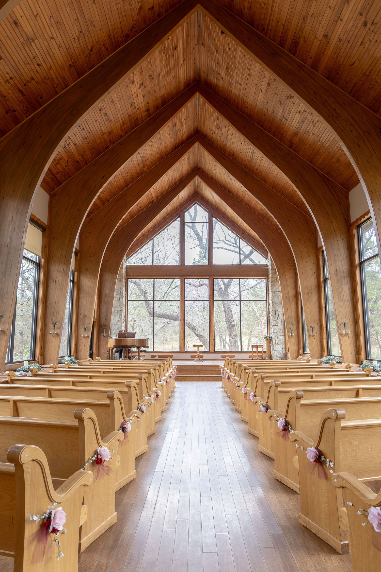 Elegant wooden chapel interior with arched ceilings, light wood pews adorned with floral decorations, and large windows overlooking trees.