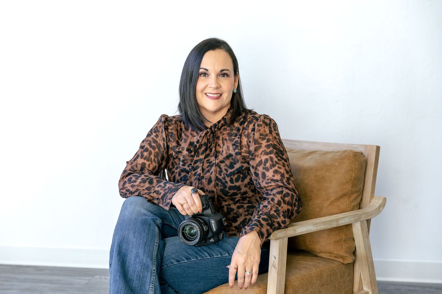 Becky Shea, wedding photographer, in a brown top and jeans, sits casually in a wooden chair against a white wall, posing with a camera.