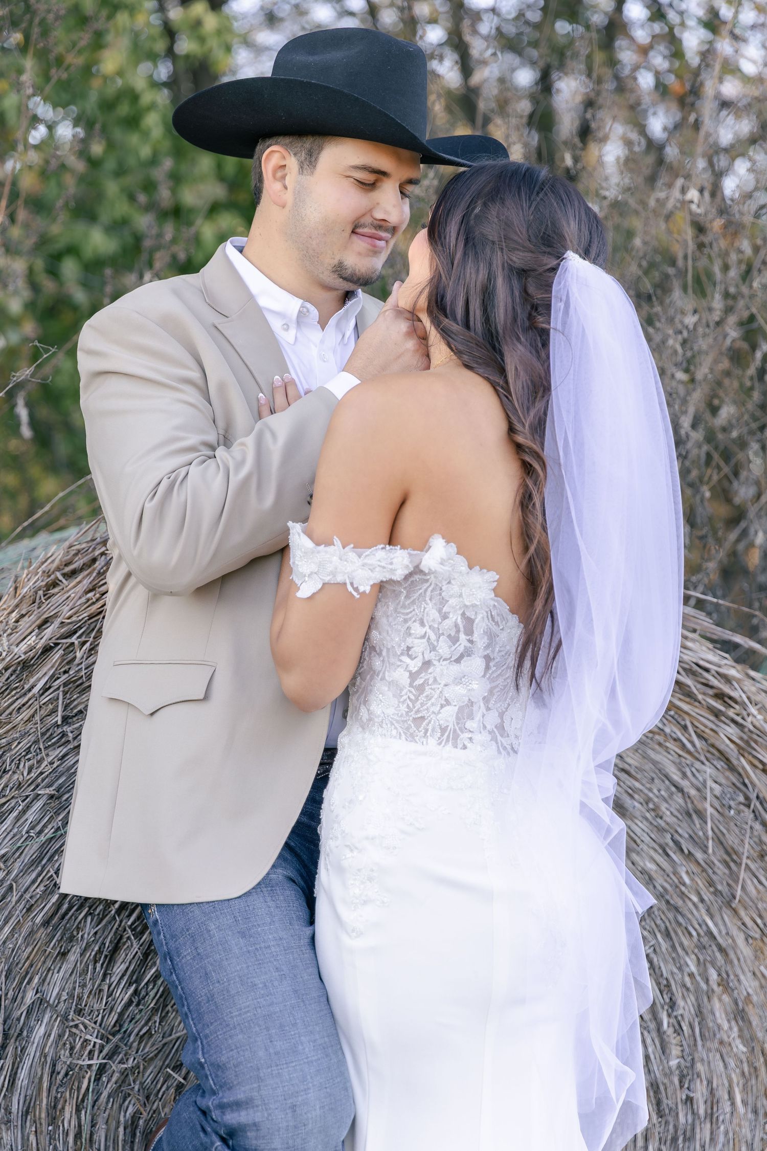 Wedding couple embraces in front of rustic hay bales wearing western-inspired attire.
