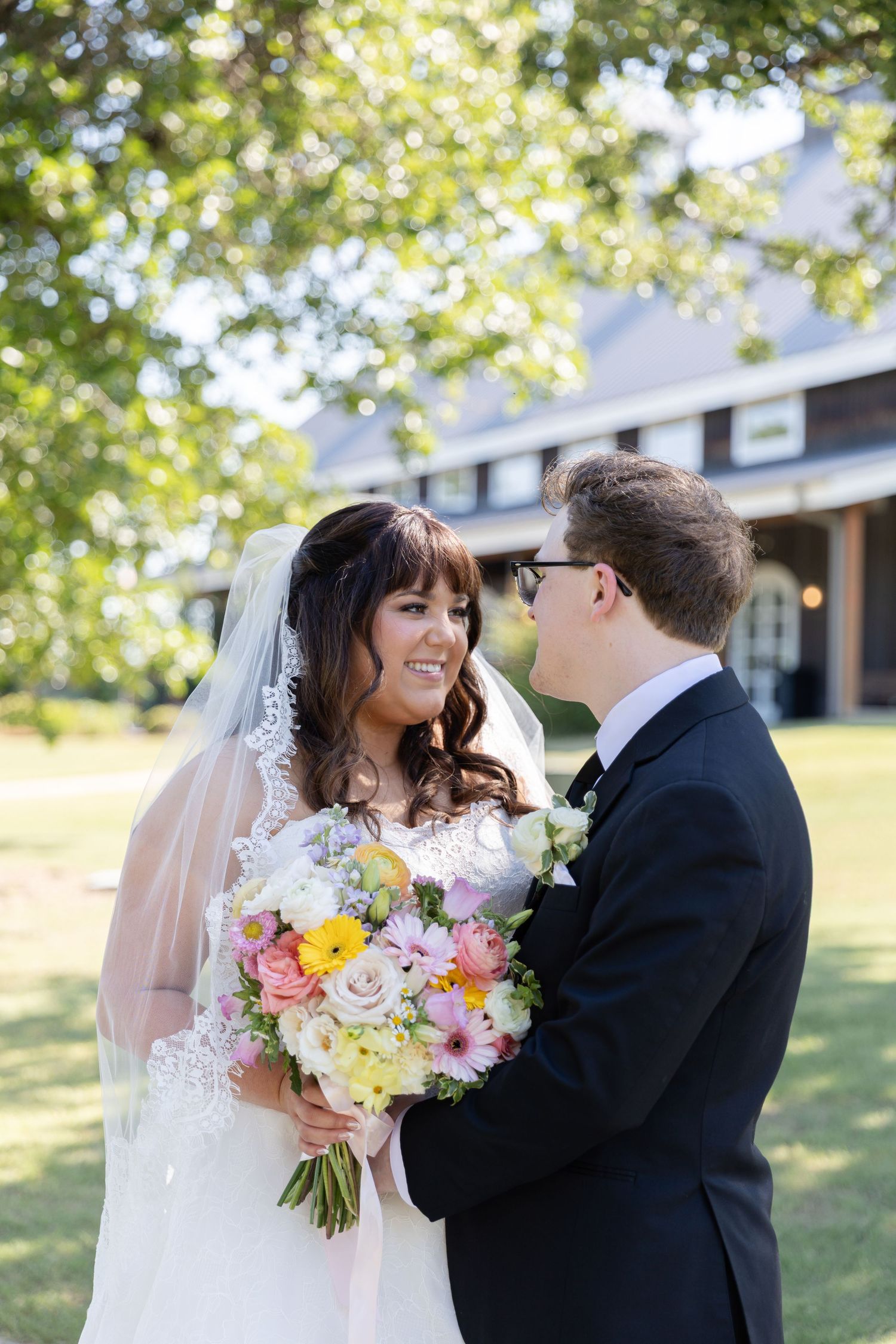Bride and groom share a romantic moment outdoors, bride holding colorful bouquet in white lace gown with veil.