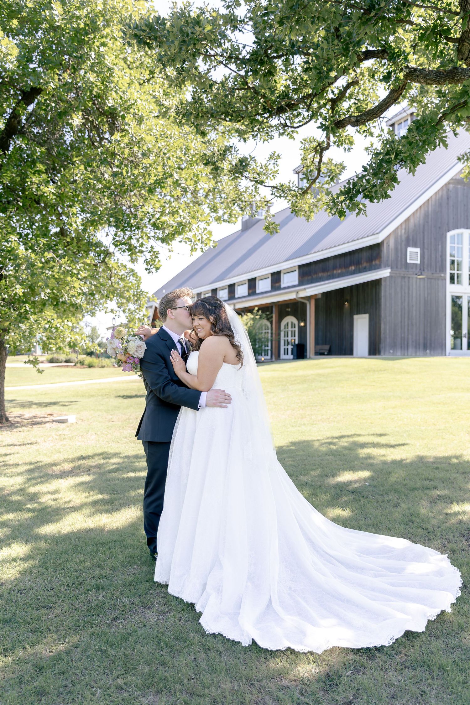 Bride and groom share a romantic embrace on green lawn beside rustic barn venue under oak tree.