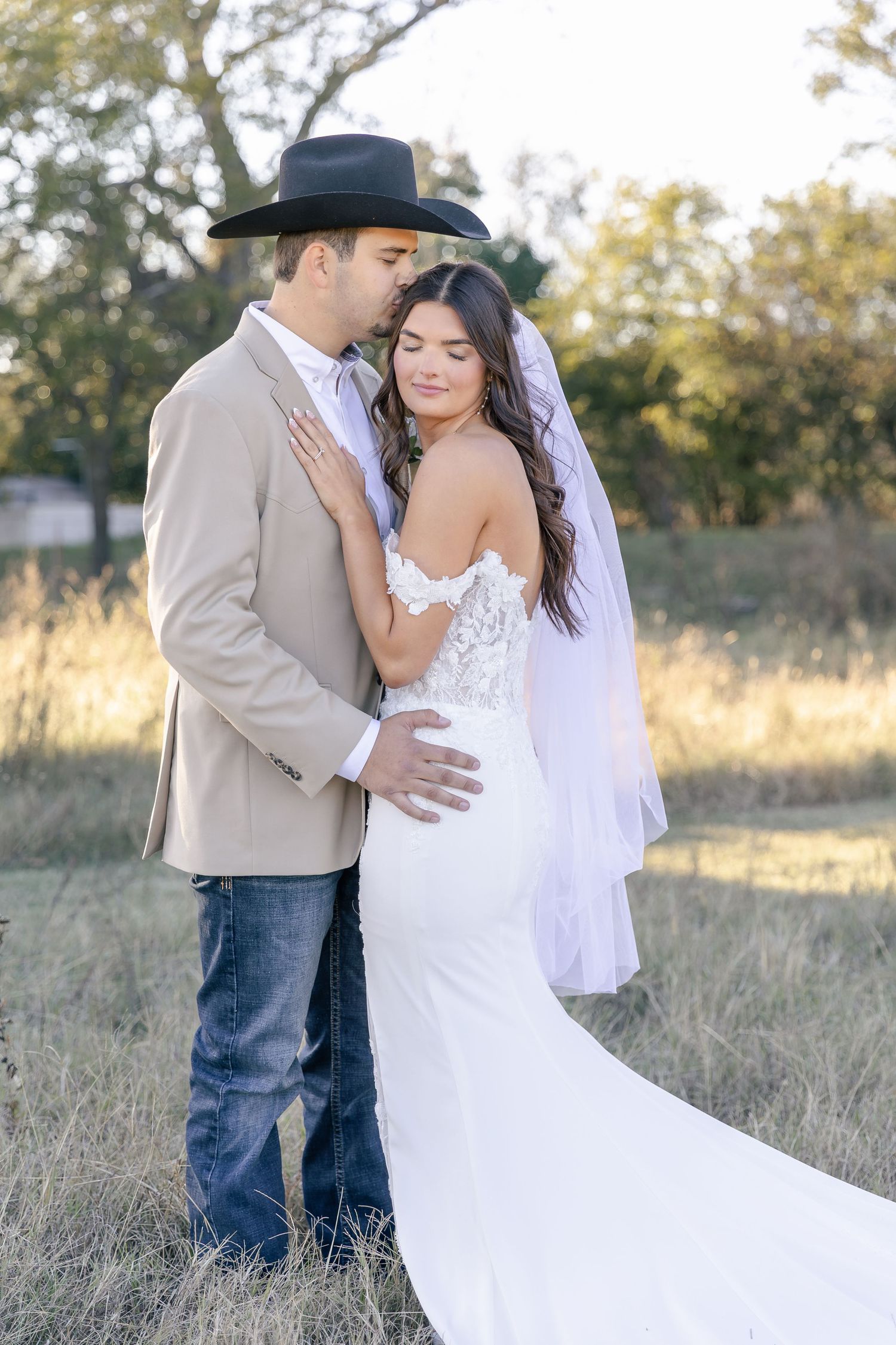 A romantic outdoor photoshoot in a sunlit field shows a couple embracing in wedding attire.