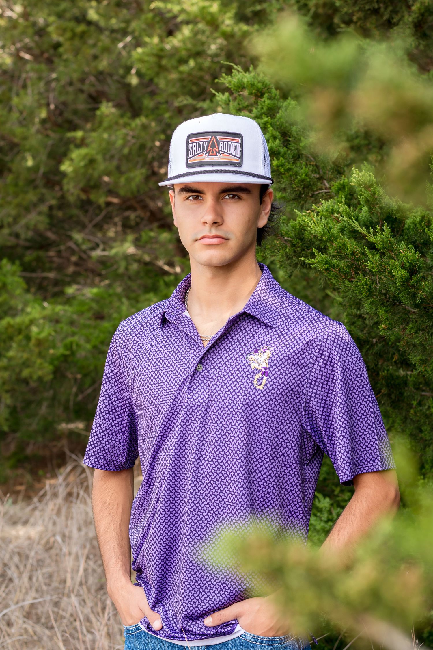 Athletic senior portrait in a purple polka dot polo and a baseball cap among greenery.
