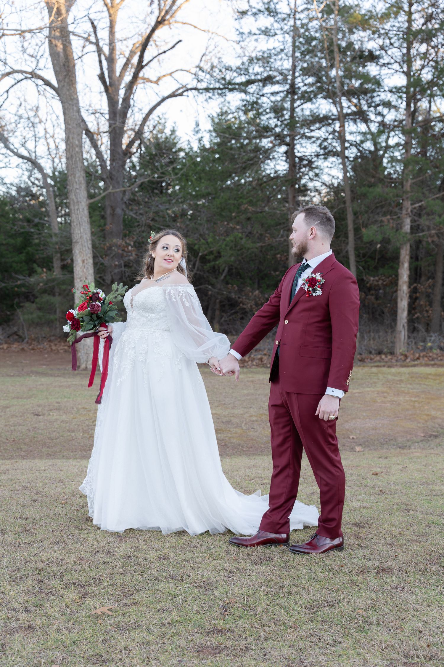 Bride in white gown and groom in burgundy suit hold hands outdoors at a winter wedding ceremony.