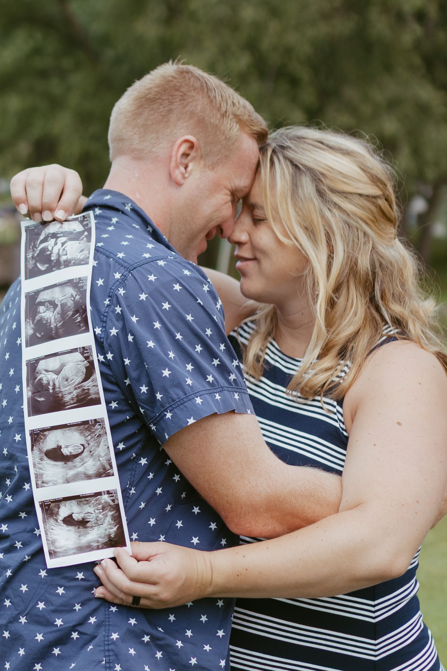 Happy couple embracing outdoors holding ultrasound photos, announcing pregnancy with foreheads touching in a tender moment.