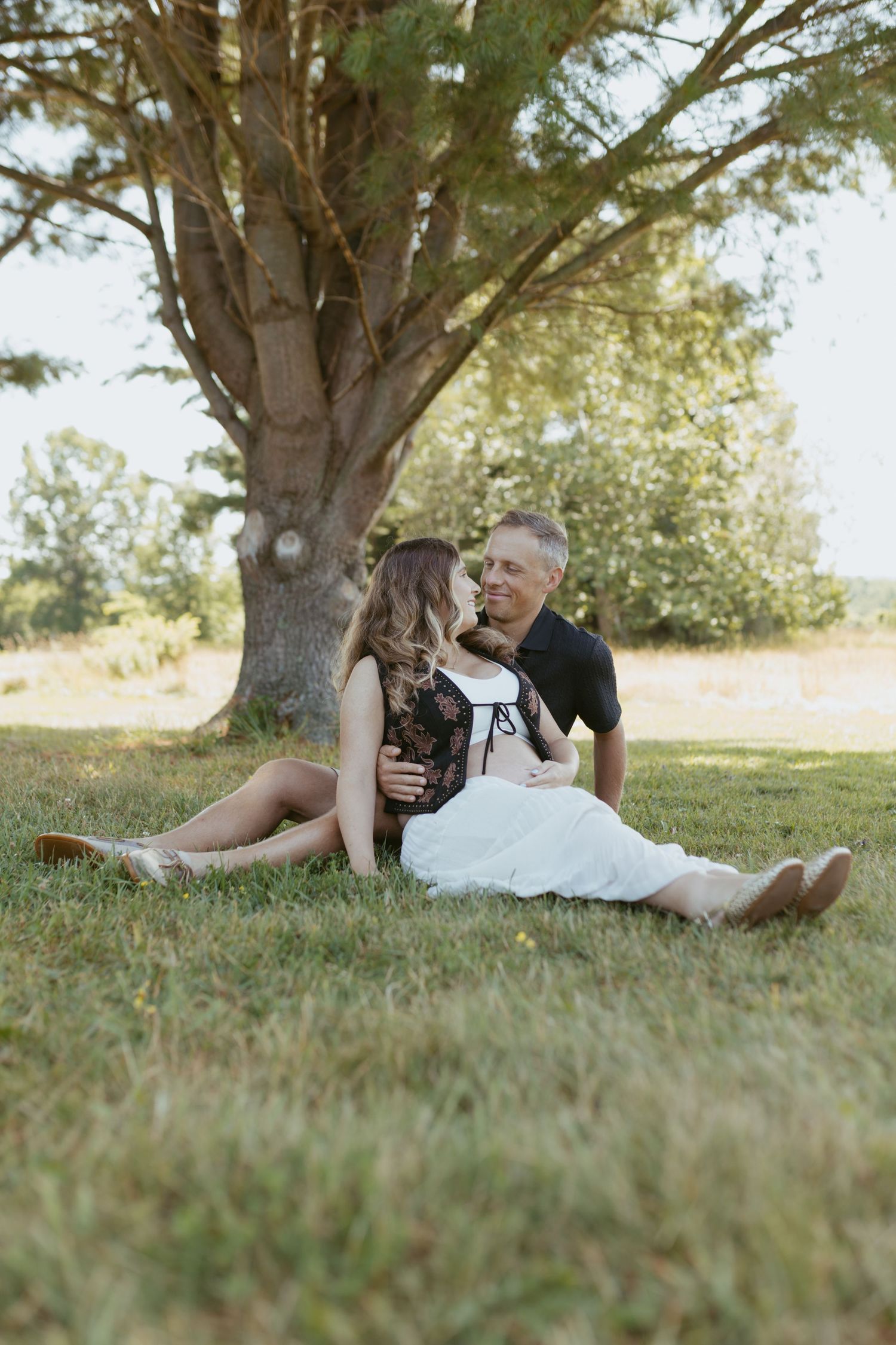 Couple sitting together on grass under a large tree, sharing a romantic moment in a sunny outdoor setting.