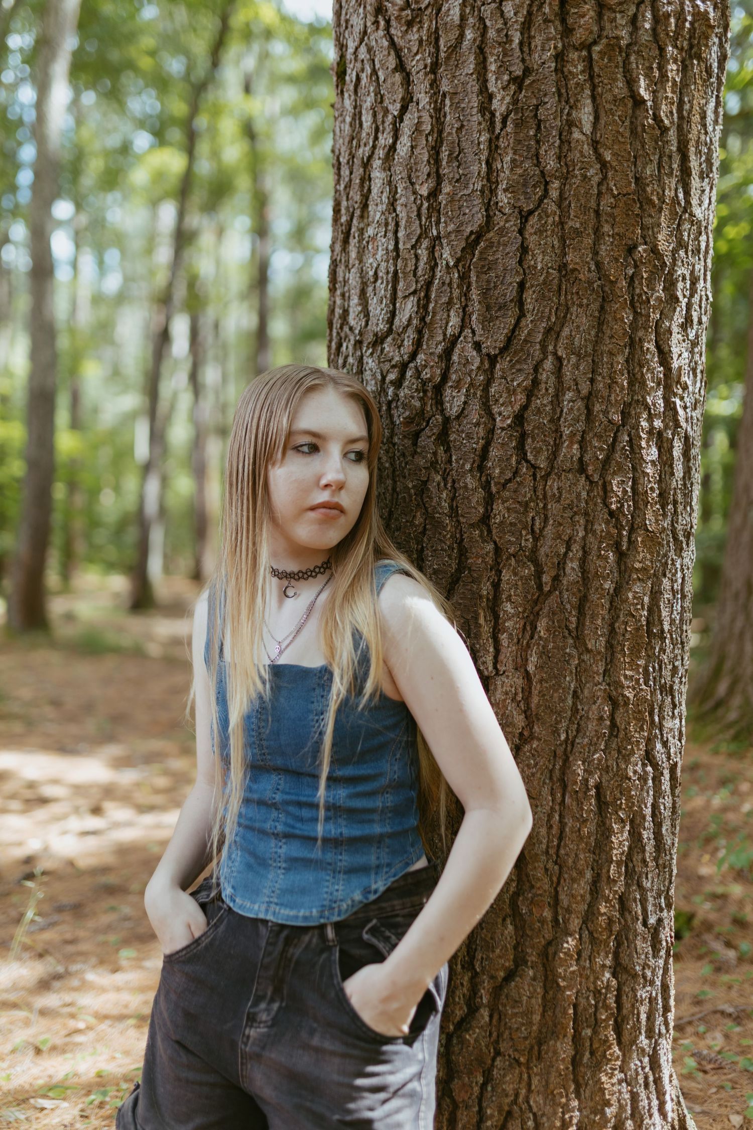 Young woman in denim top and black jeans leaning against a tree trunk in a sunlit forest.