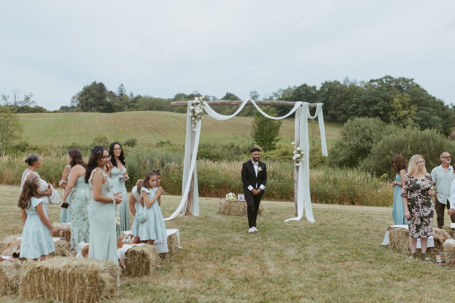 Outdoor wedding ceremony on a green hillside with bridesmaids in mint dresses, groom at floral arch with white draping.