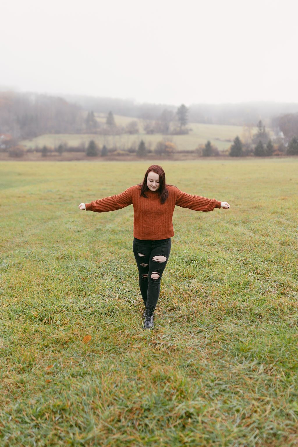 Person in rust sweater and ripped jeans spinning joyfully in a misty field.