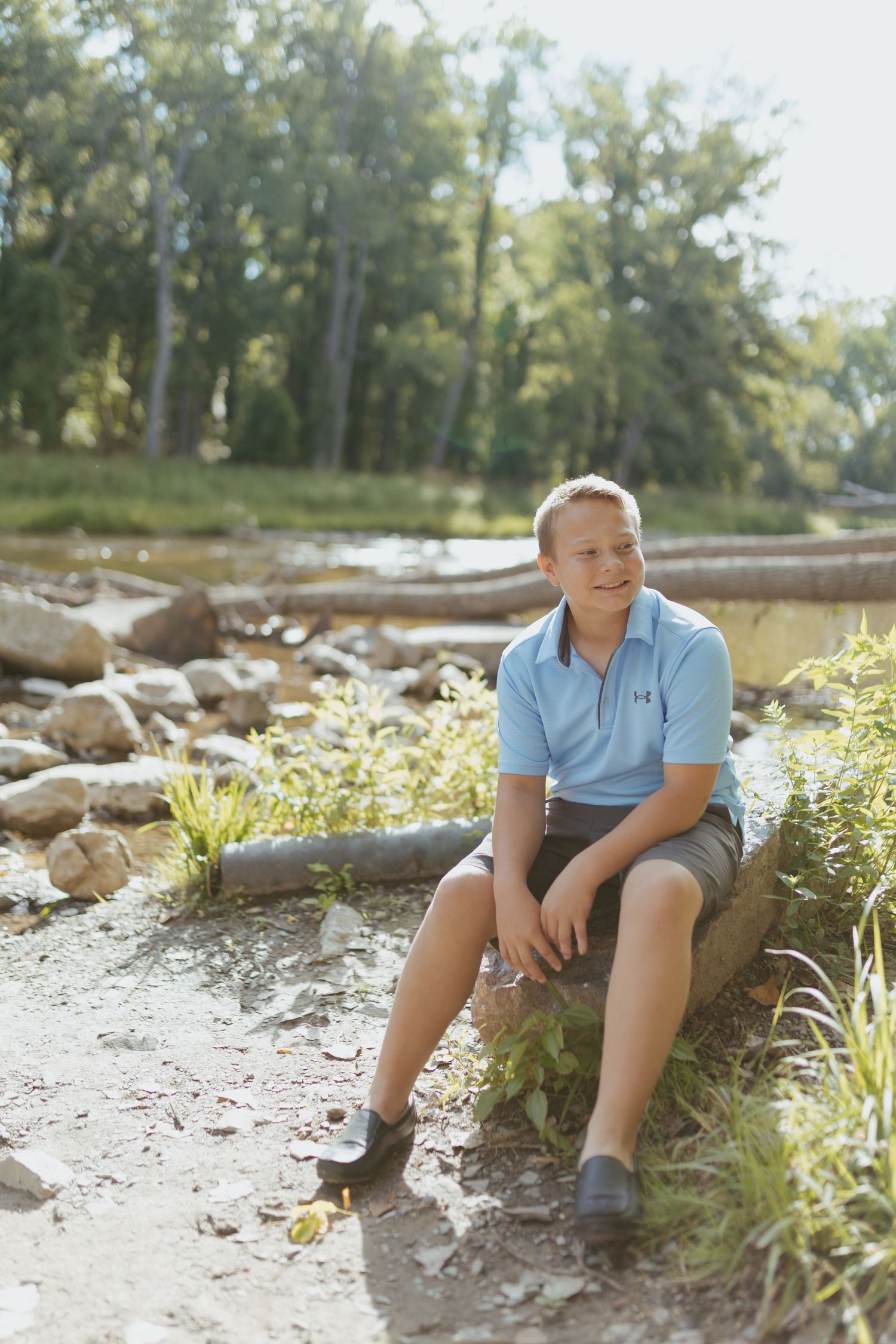 A smiling teenage boy in a blue polo shirt and shorts sits on a log near a rocky riverbank surrounded by trees.