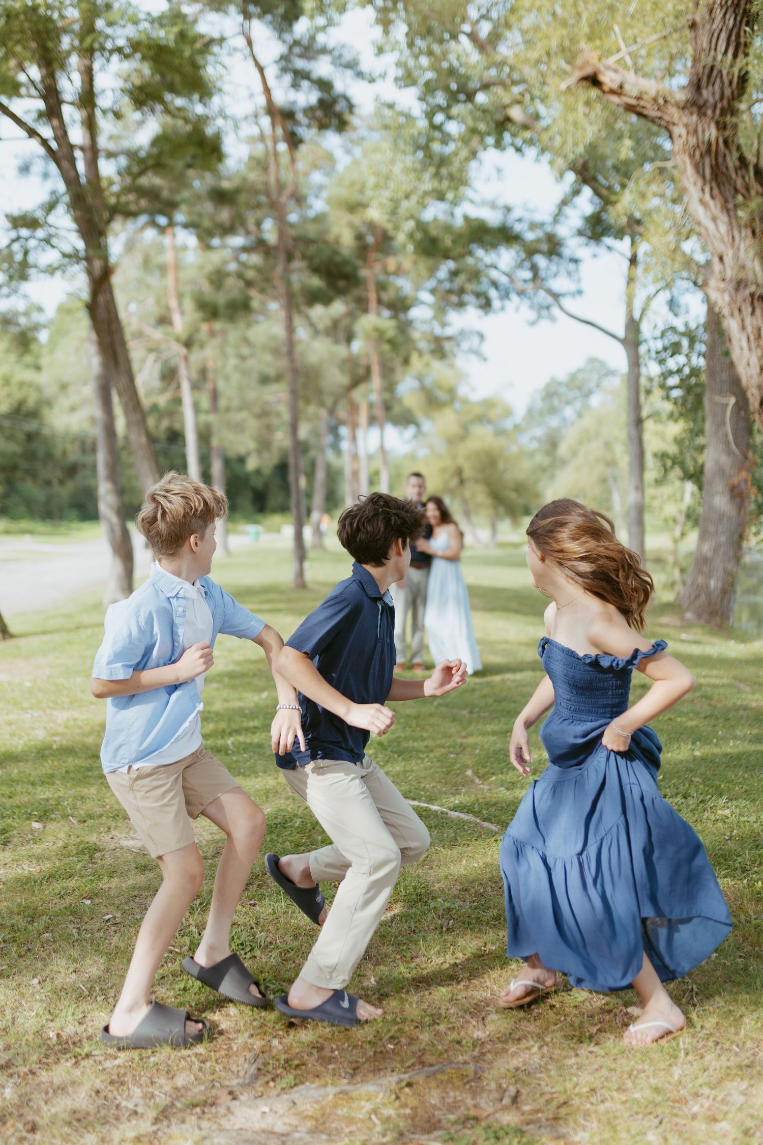 Three children playing and dancing outdoors in a sunny park, with two adults standing among tall trees in the background.