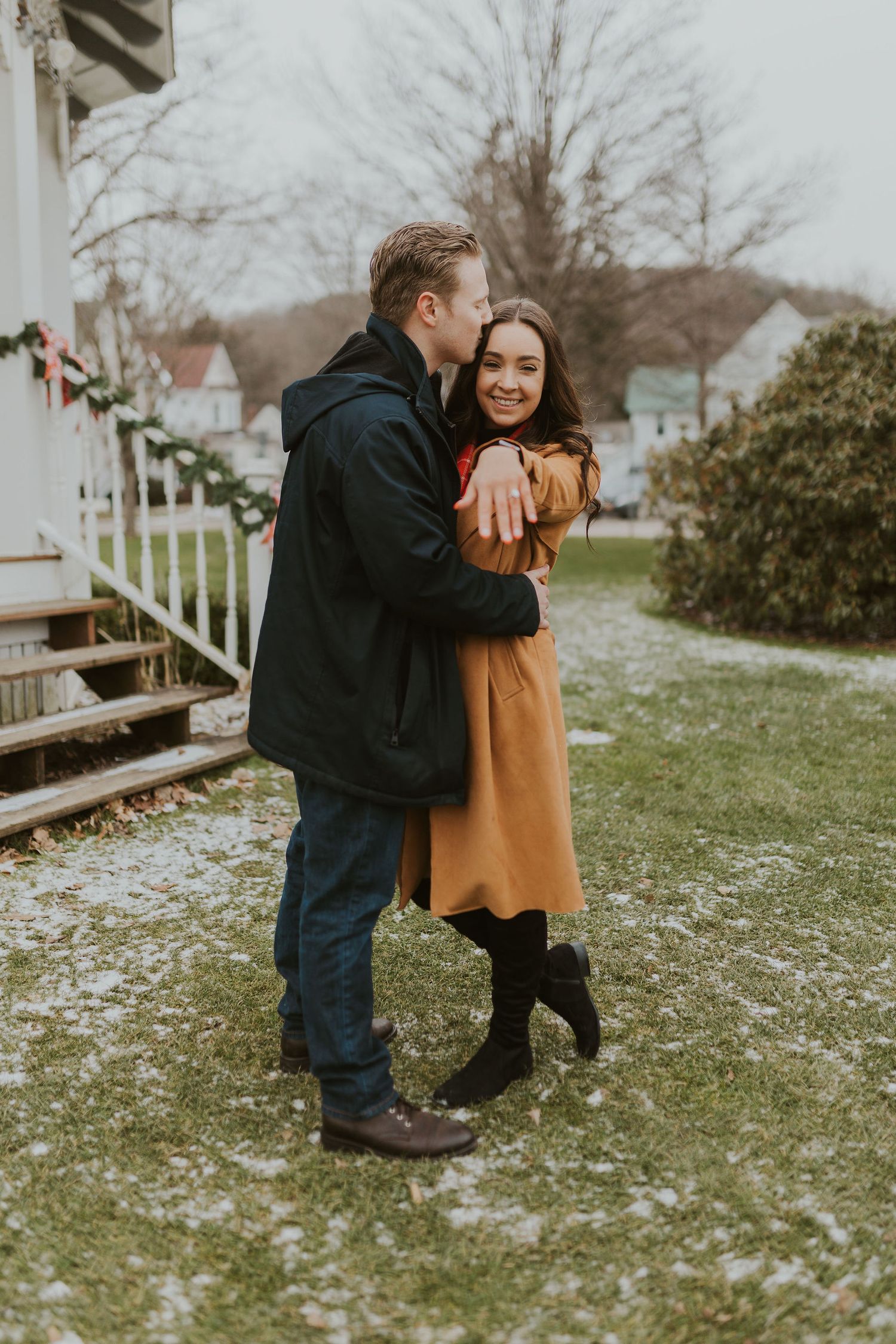 Couple embracing outdoors in winter, man kissing woman's cheek near decorated porch.