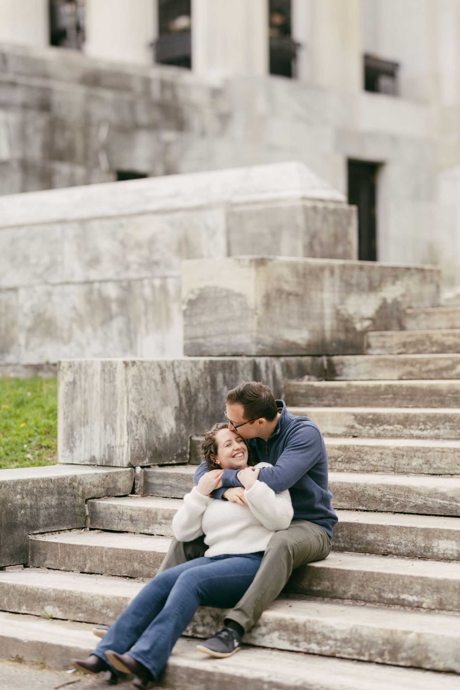 Couple sitting on stone steps, man kissing woman's cheek while embracing her outdoors near a historic building.