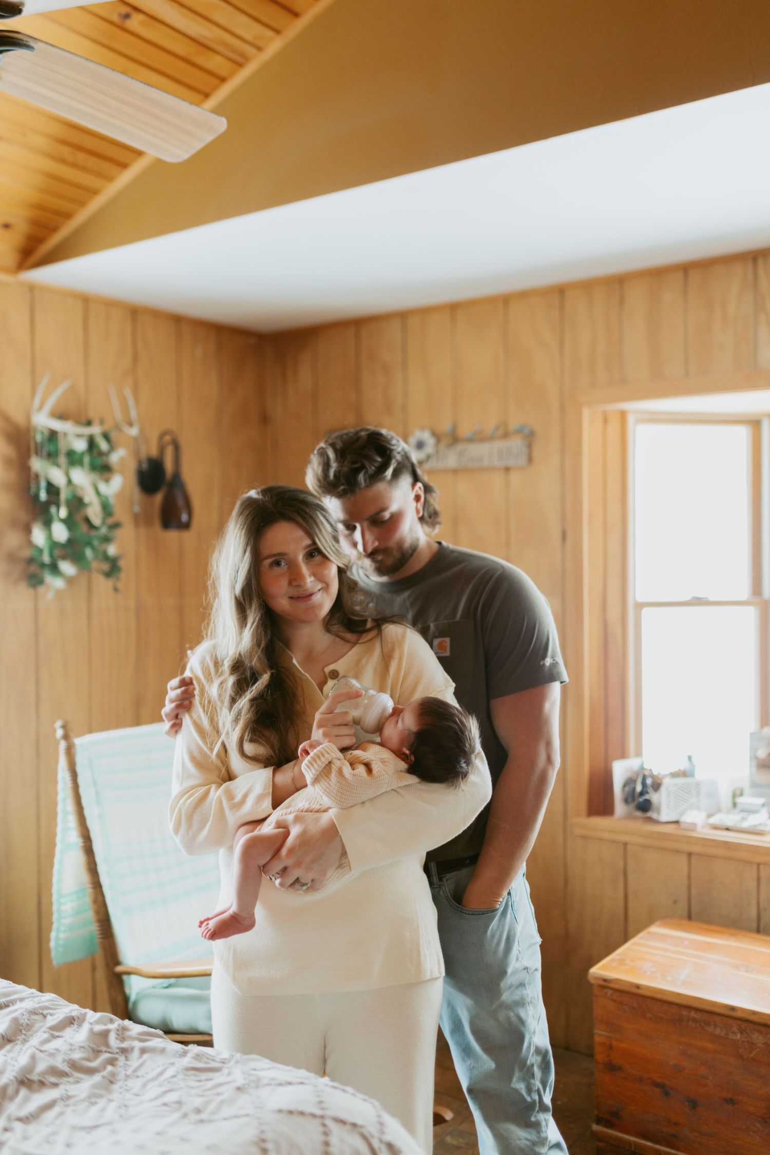 Young couple holding newborn baby in rustic wood-paneled bedroom with warm lighting.