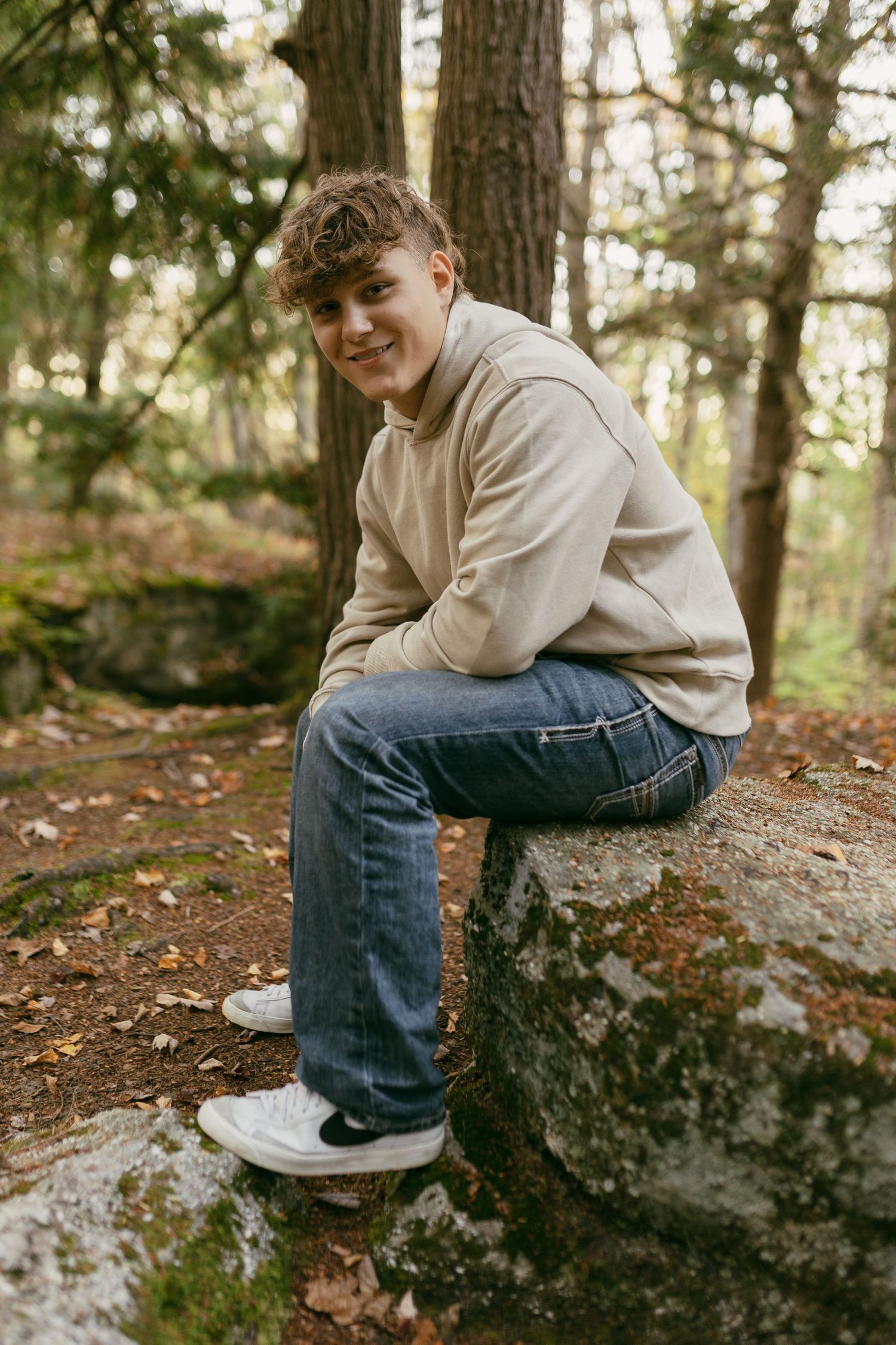 Smiling teen boy in beige hoodie and jeans sitting on mossy rock in forest.