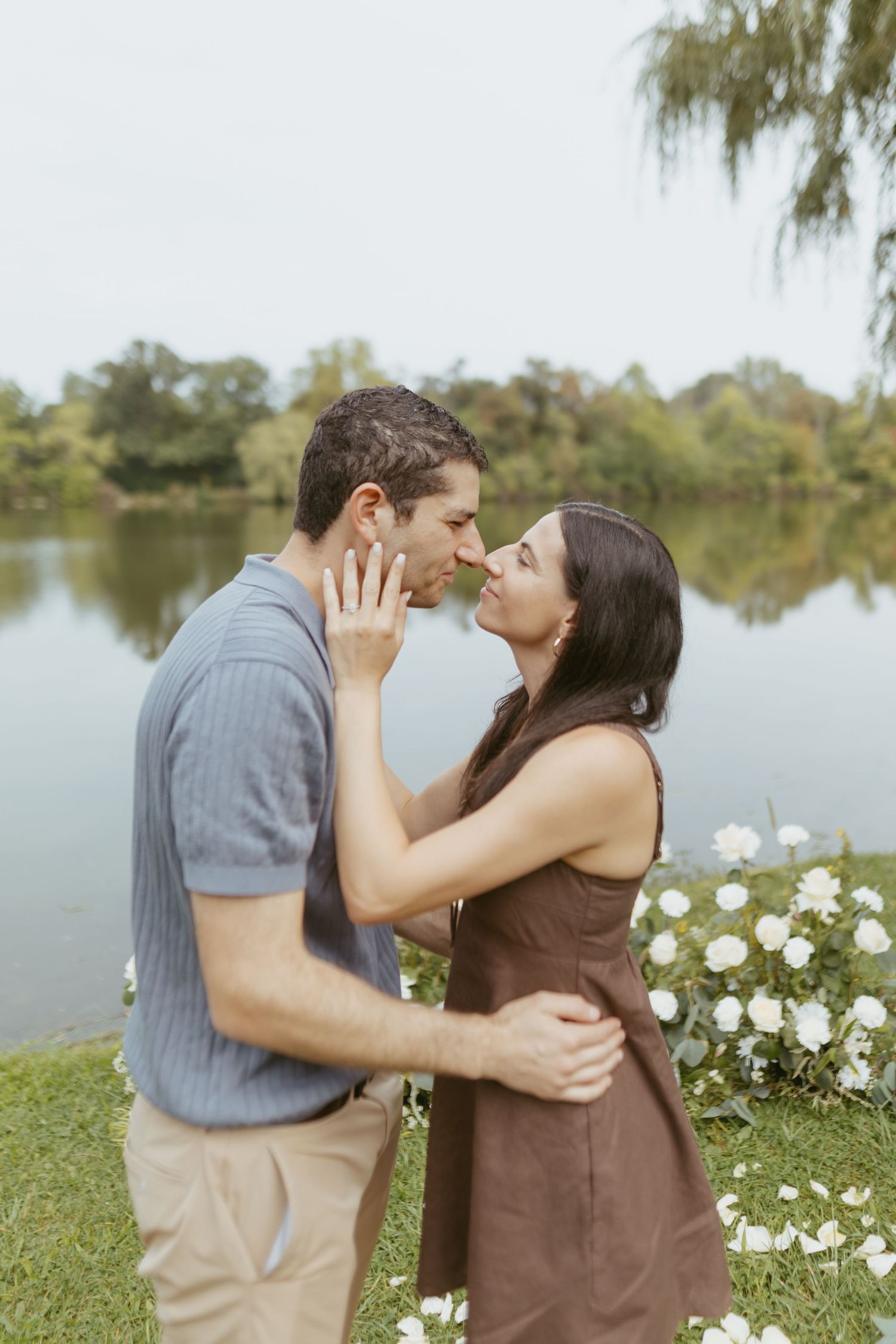 Couple sharing a romantic moment by a serene lake, surrounded by white flowers and lush greenery.
