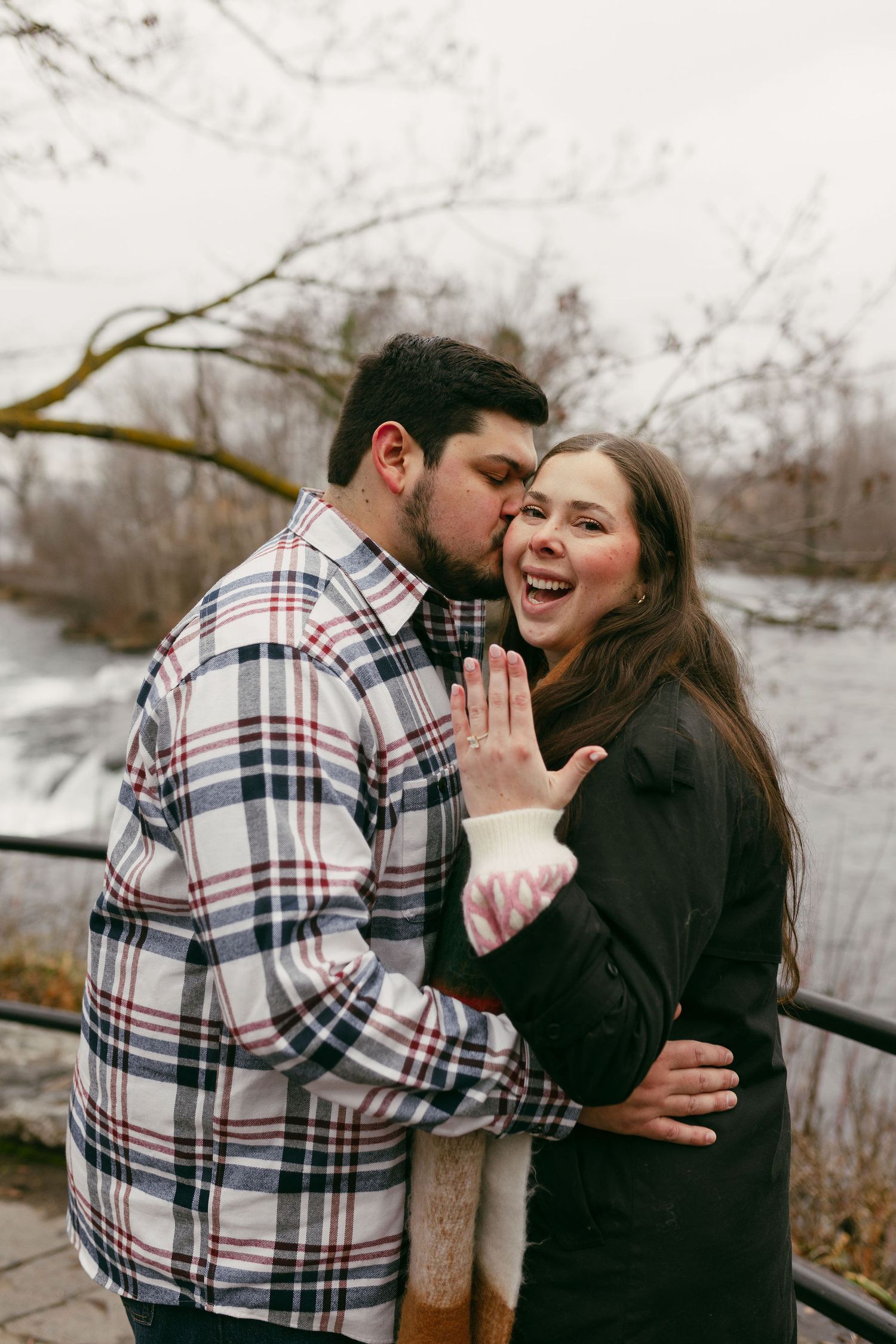Couple celebrating engagement outdoors, woman showing ring while man kisses her cheek, laughing joyfully near a winter river.