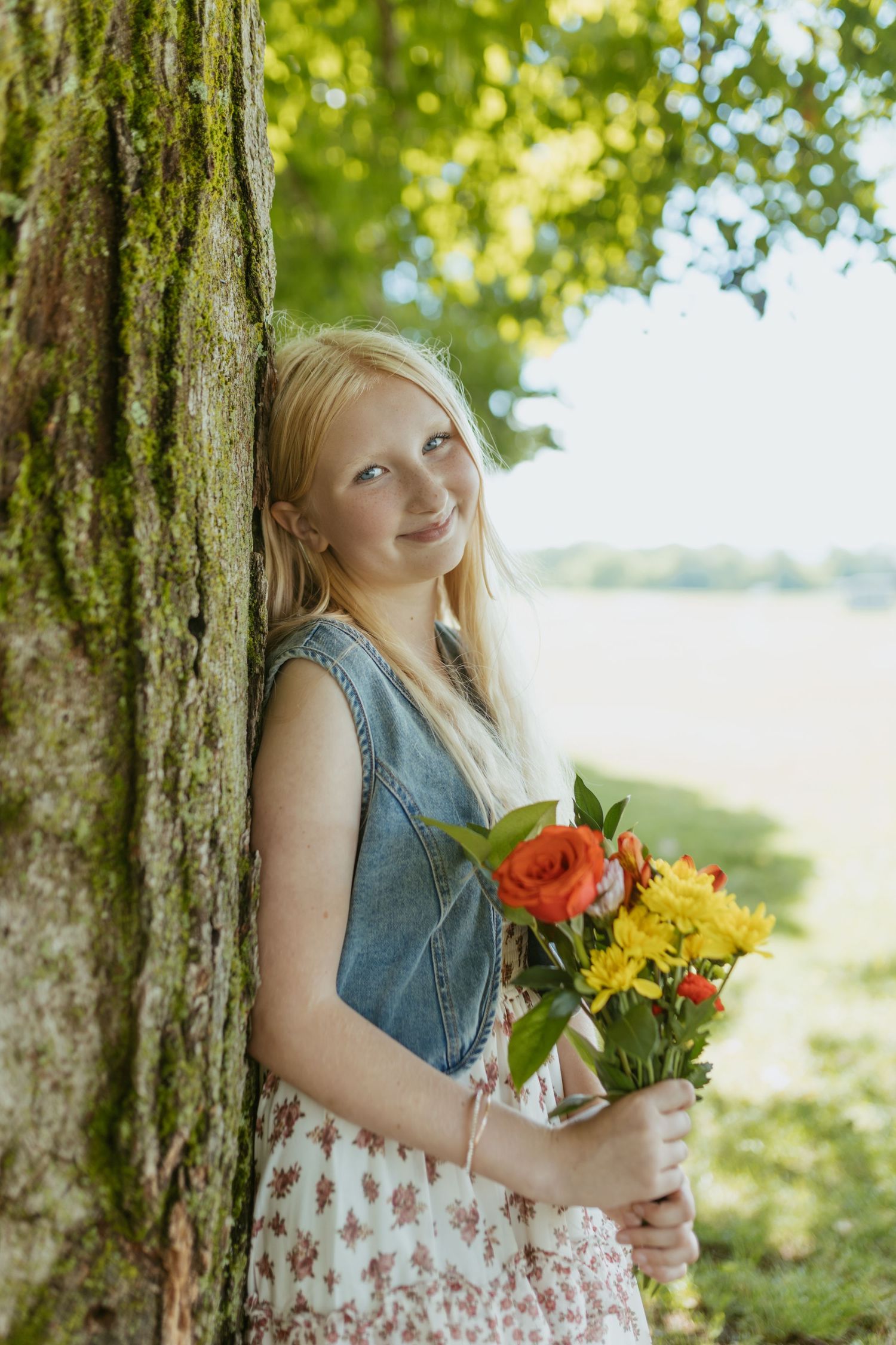 Smiling blonde girl leaning against a mossy tree, holding a bouquet of orange and yellow flowers outdoors.
