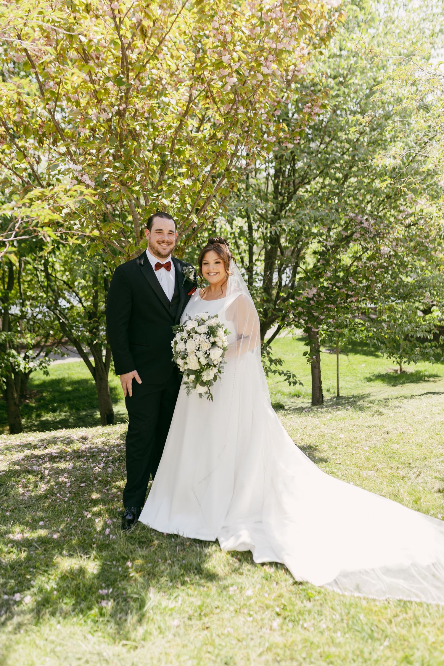 Bride and groom posing together outdoors among blooming trees, bride holding white floral bouquet.