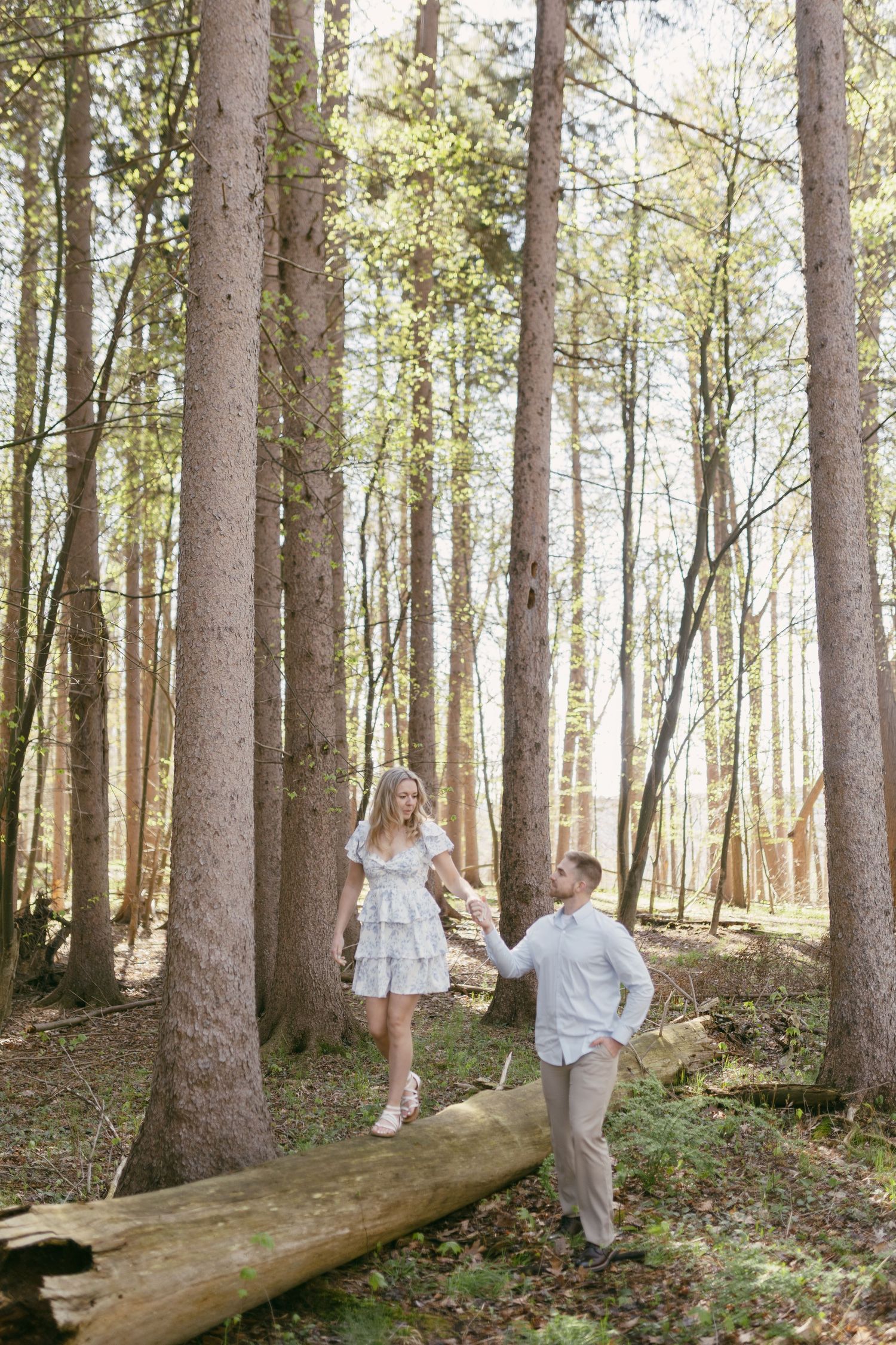 Couple holding hands and walking through a sunlit forest, woman in white dress balancing on a fallen log.