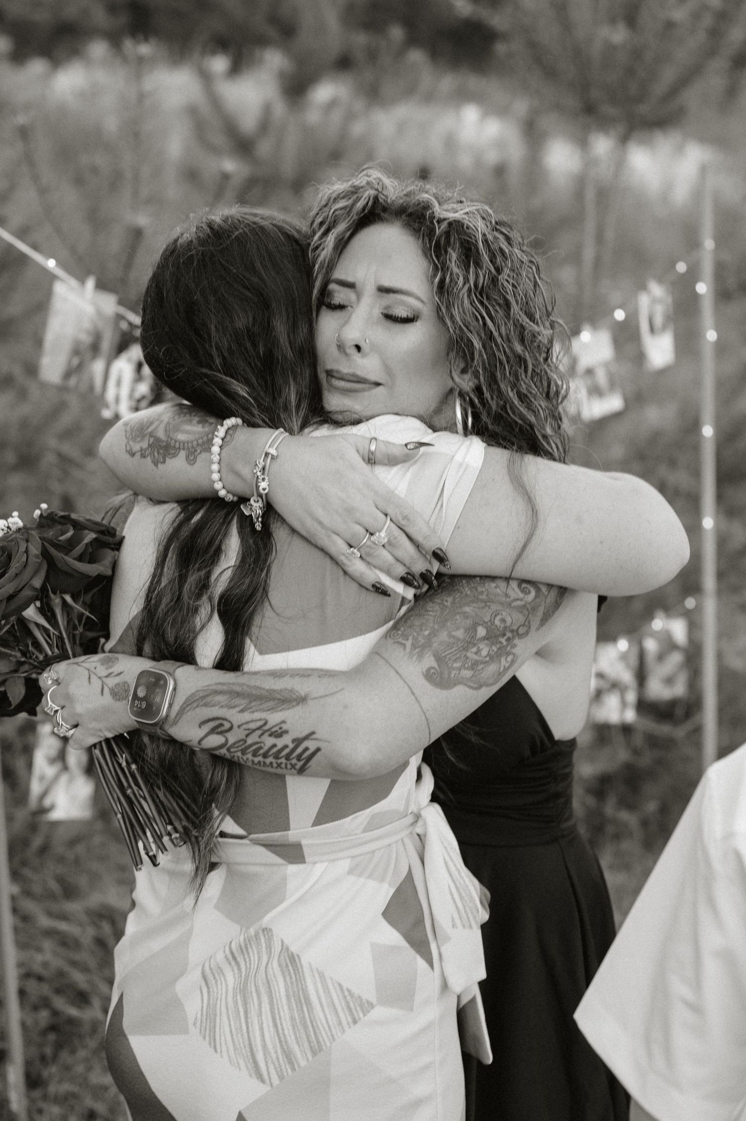 Two women share an emotional hug at an outdoor wedding, one in bridal attire, both with tattoos, in black and white.