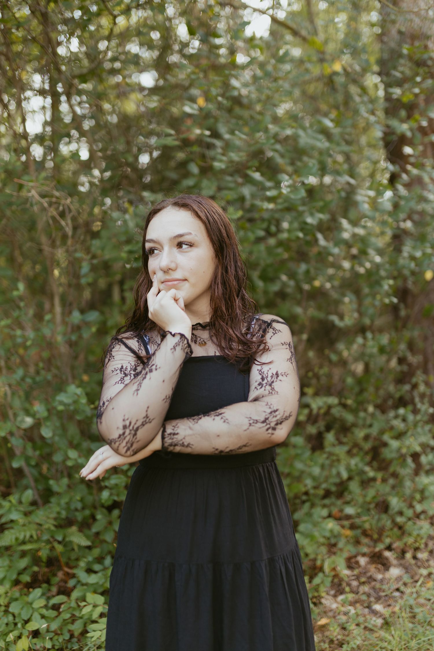 Young woman in black lace dress posing thoughtfully outdoors surrounded by lush green foliage.