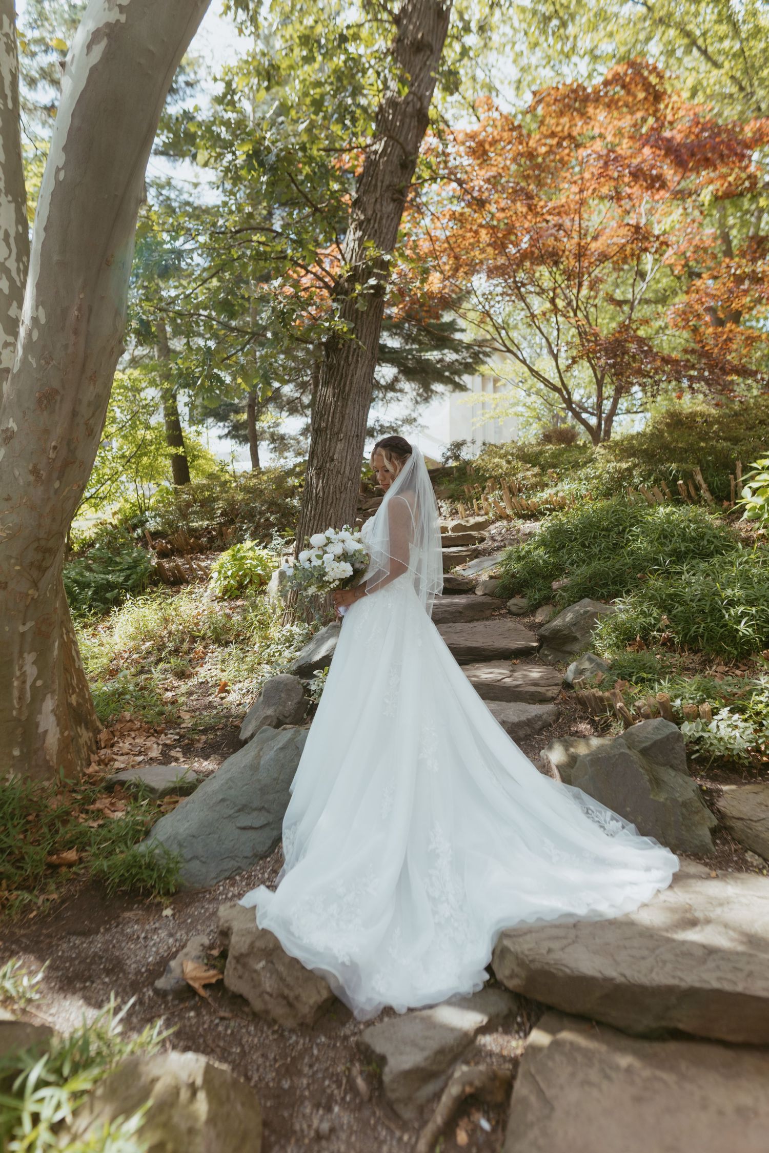 Bride in a flowing white gown and veil stands on stone steps surrounded by lush garden trees holding a bouquet.