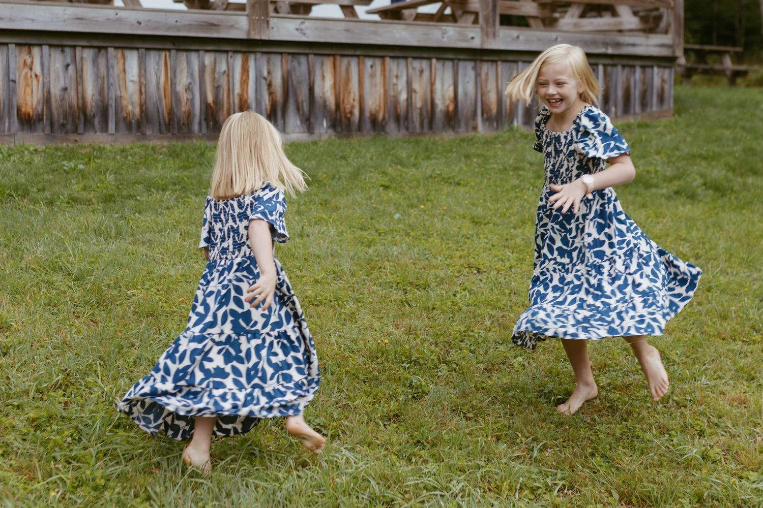 Two young girls in matching blue floral dresses twirling and playing barefoot on green grass outdoors.