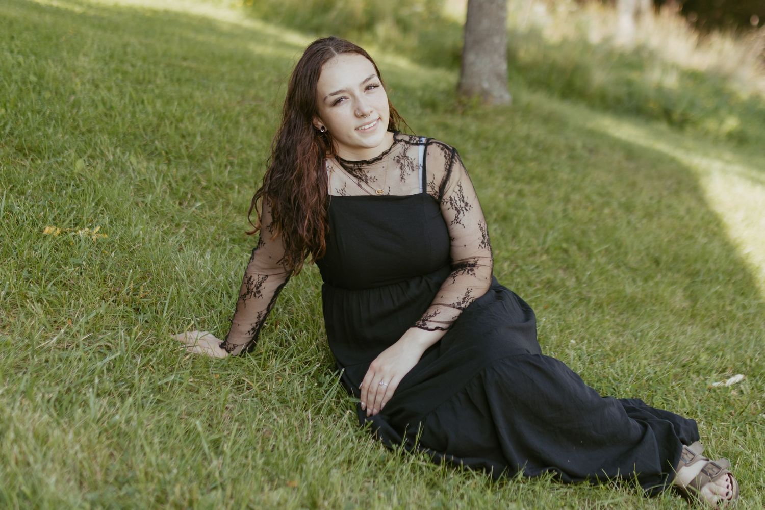 Young woman in black dress sitting on green grass in a park, smiling gracefully.