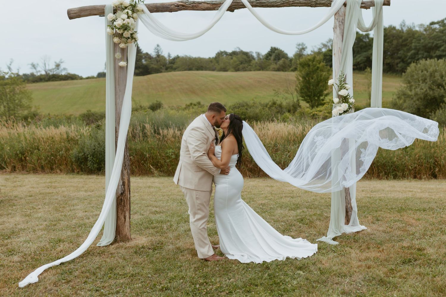 Bride and groom share a kiss under a floral arch with flowing white draping at an outdoor wedding ceremony.