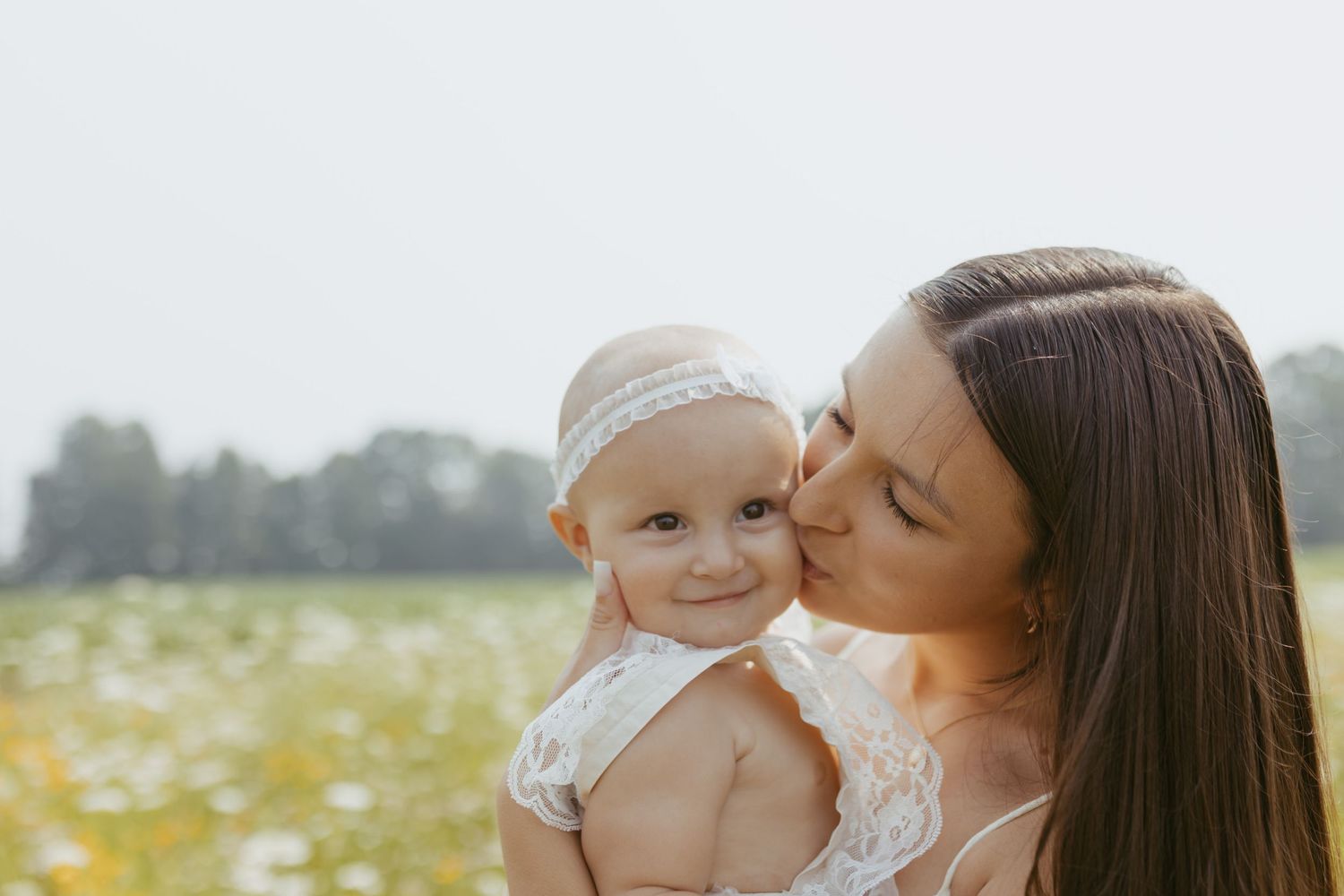 A mother tenderly kisses her smiling baby girl in a sunlit flower field.