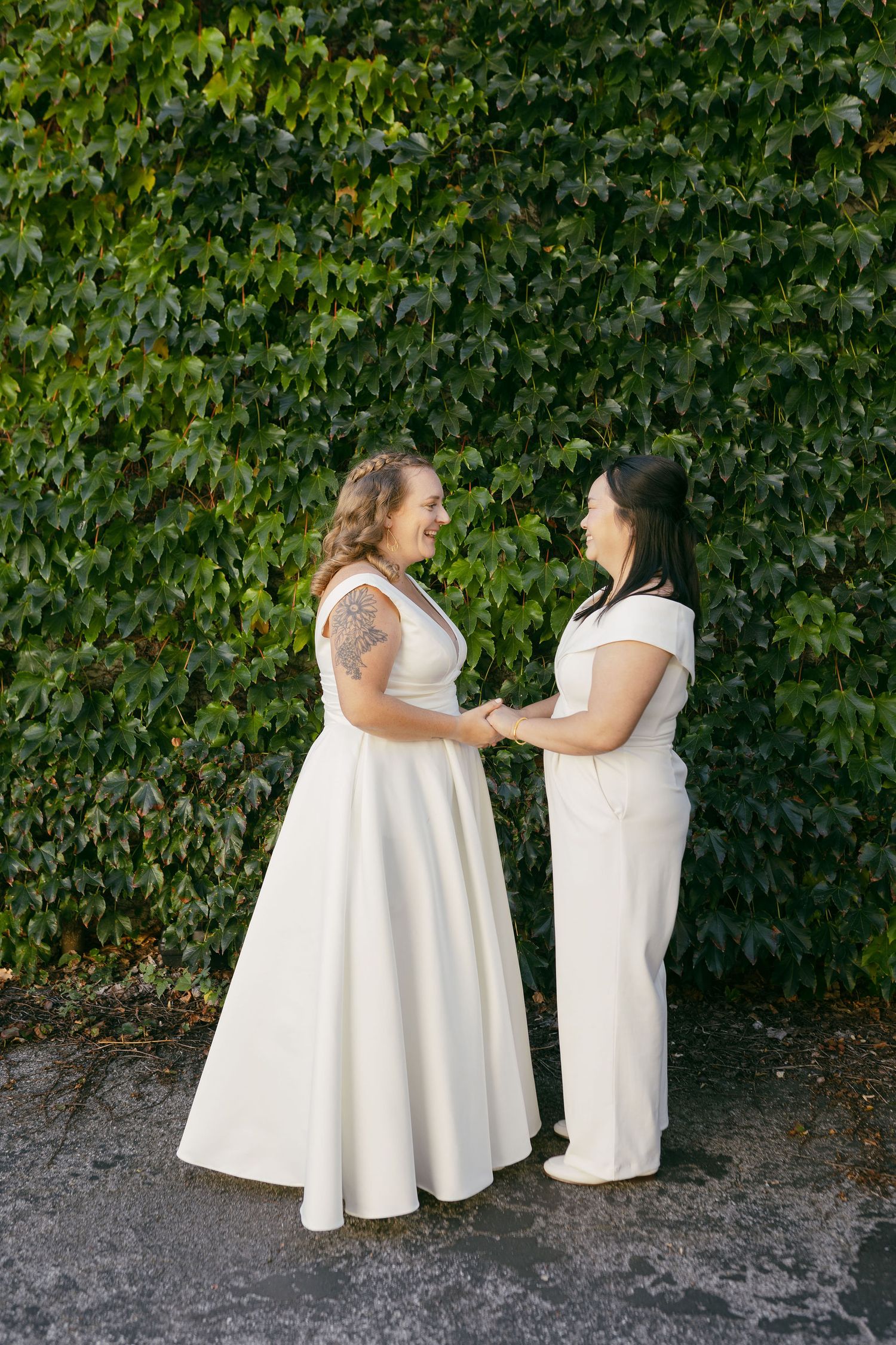 Two brides in elegant white dresses share a joyful moment against a lush ivy wall.