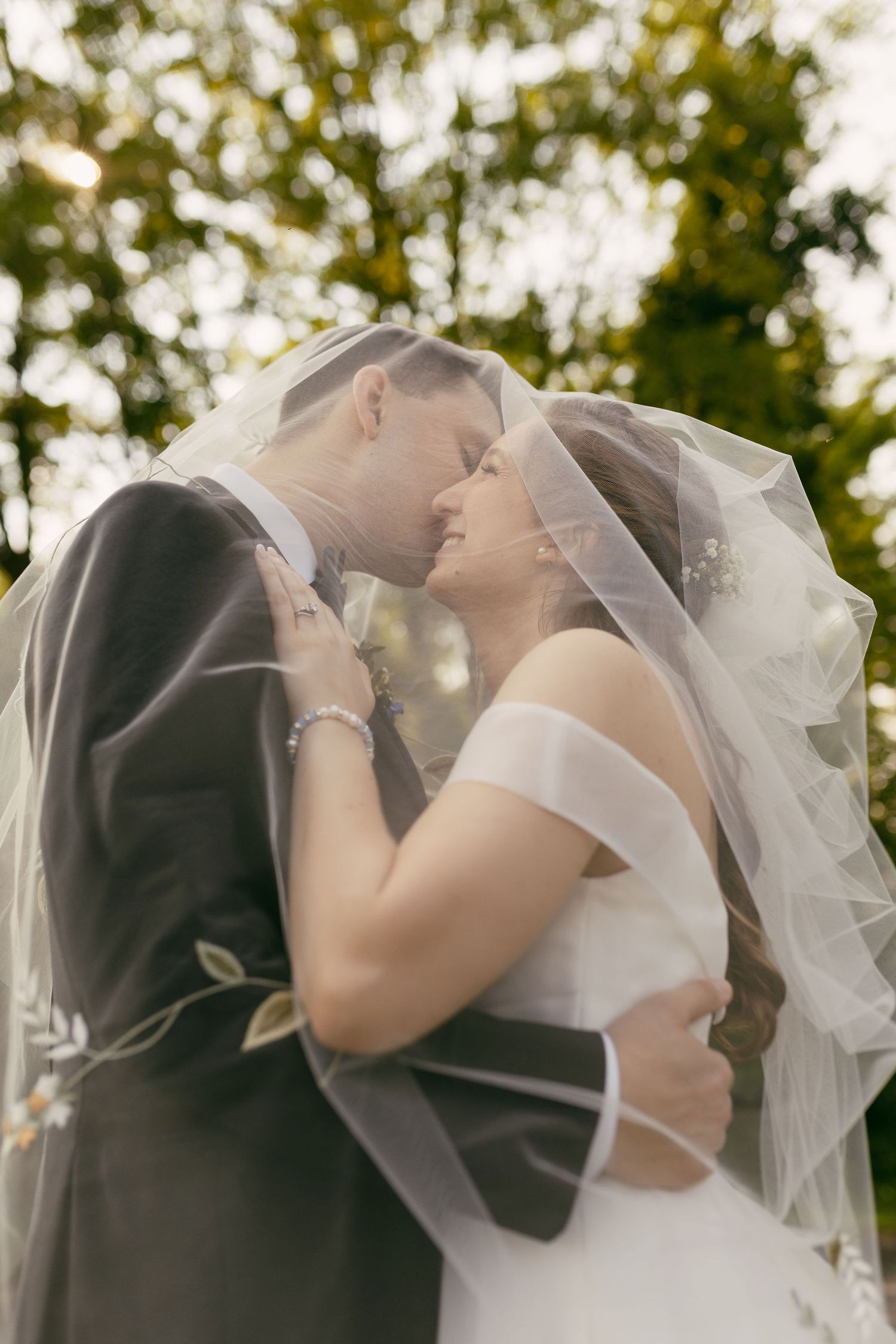 Romantic moment between newlyweds under a flowing veil with sunlit trees in background.