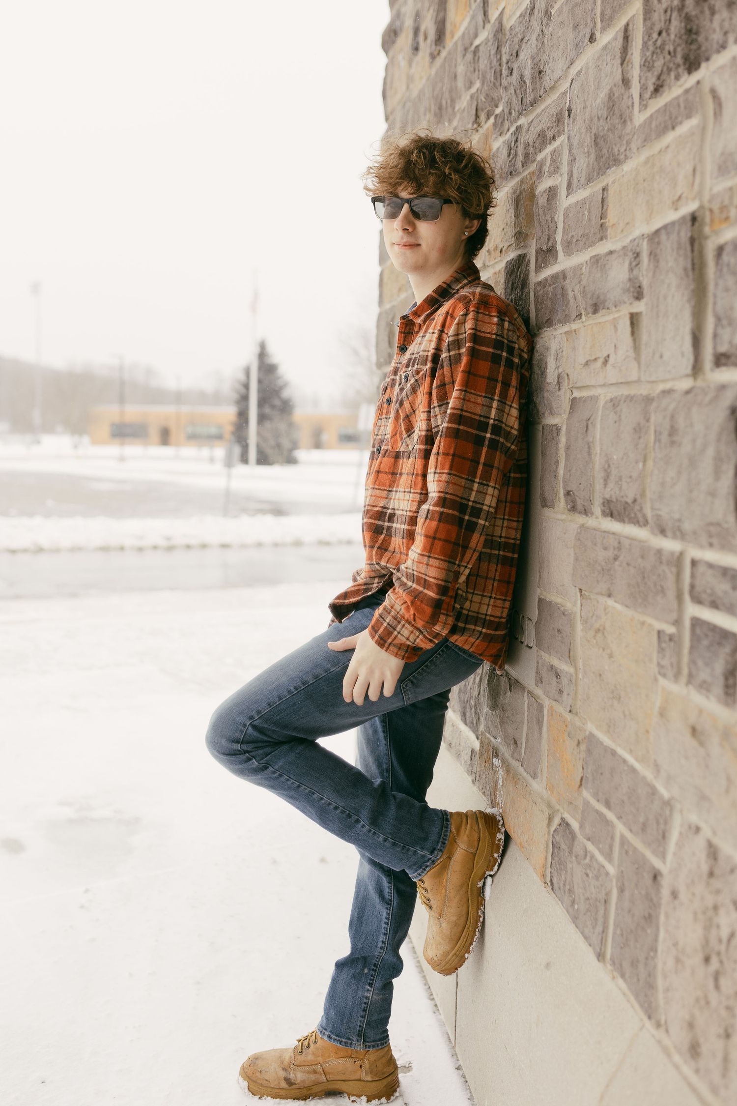 Person in plaid shirt and jeans leaning against stone wall in winter scene.