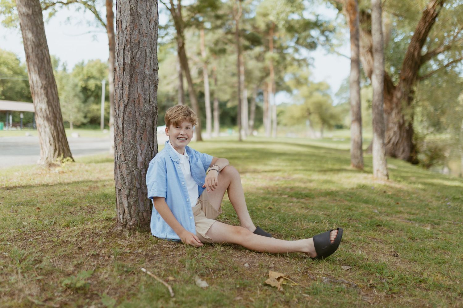 A smiling boy sitting against a tree trunk on green grass in a sunny park, wearing a blue shirt and shorts.