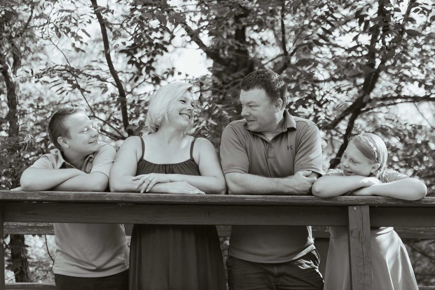 Black and white family portrait of four people leaning on a wooden railing outdoors, smiling and laughing together.