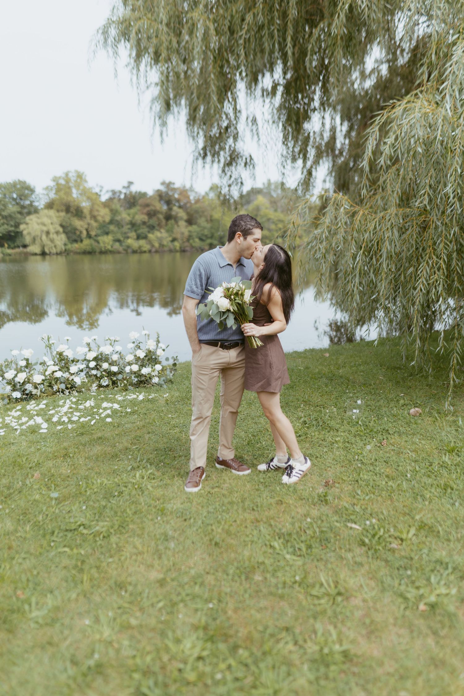 Couple sharing a romantic kiss by a serene lake, holding a white bouquet under a weeping willow tree.