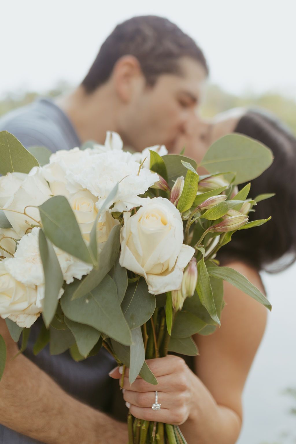 Couple kissing behind a bridal bouquet of white roses and eucalyptus, with an engagement ring visible on her hand.