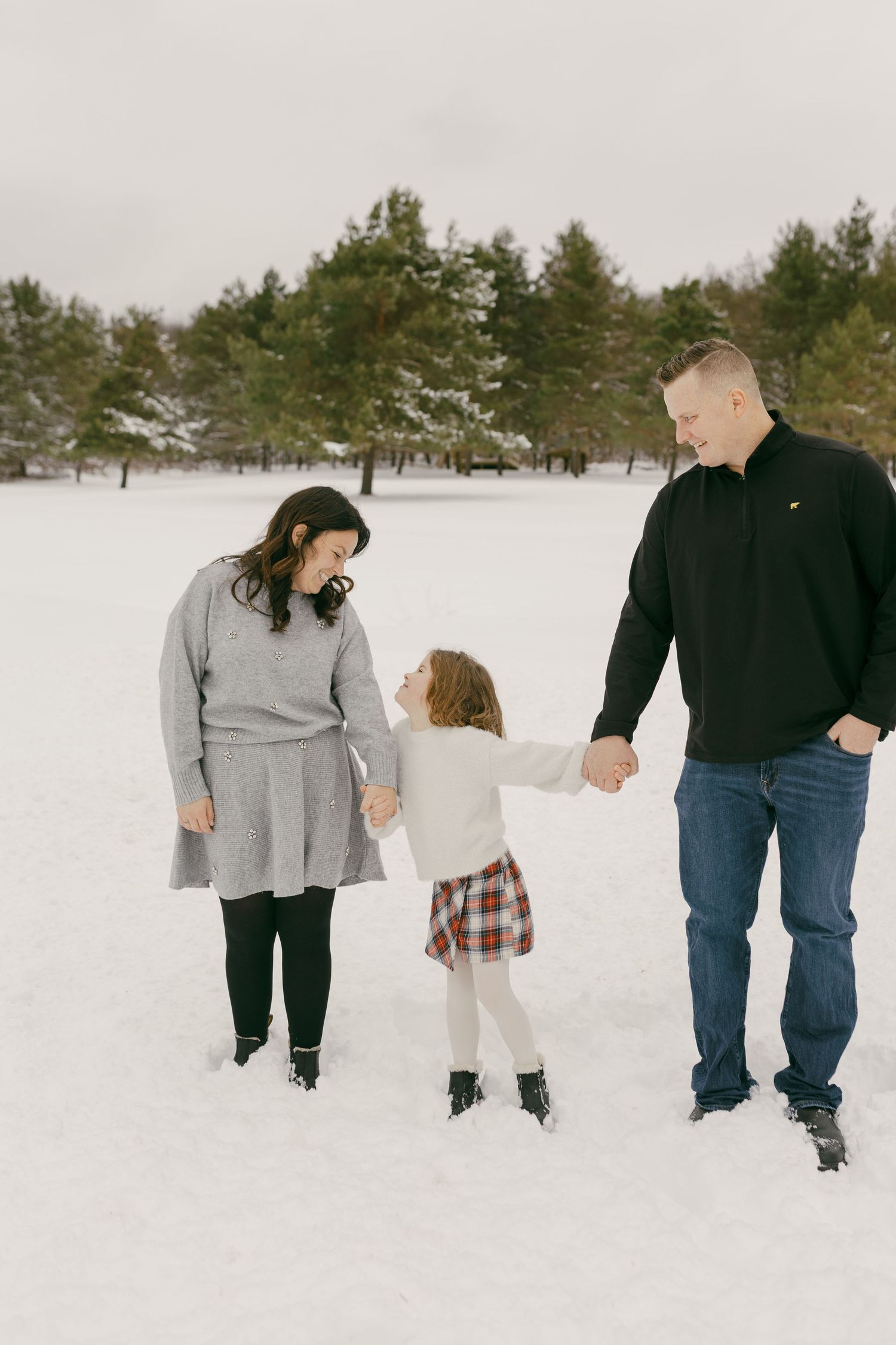 Family of three holding hands in snowy field, parents smiling at young girl in plaid skirt, evergreen trees in background.