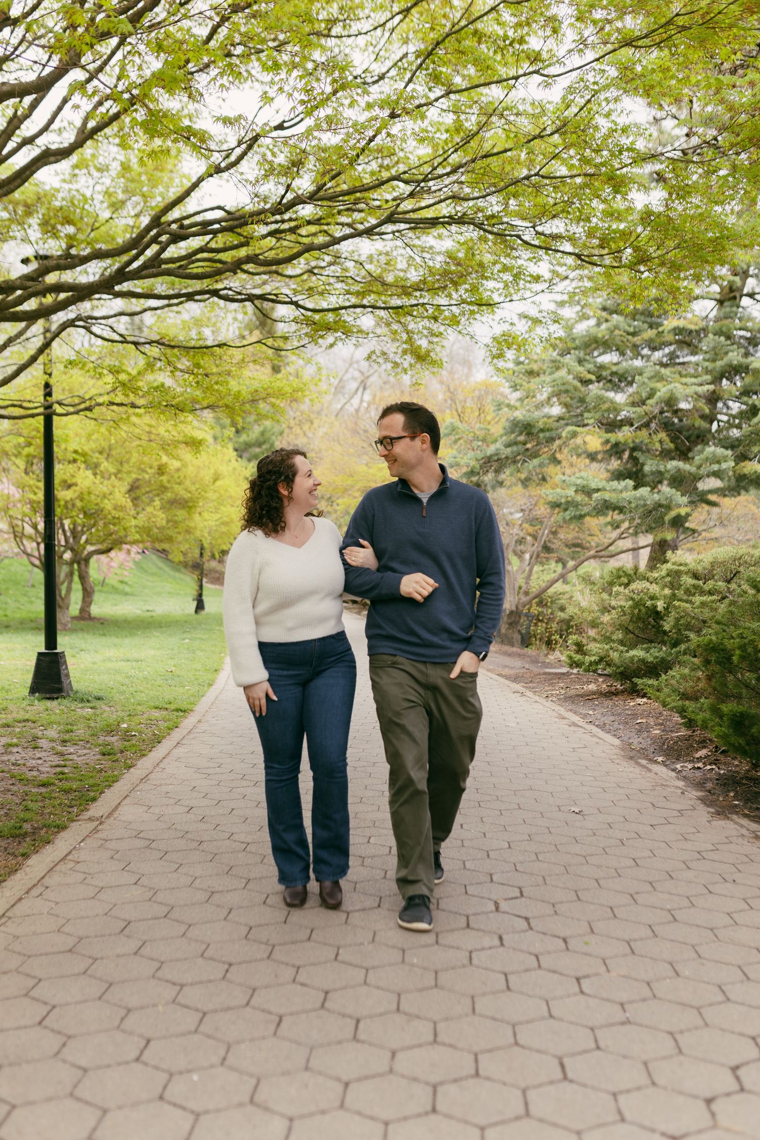 A couple walks arm-in-arm along a brick path in a lush green park, smiling at each other on a warm day.