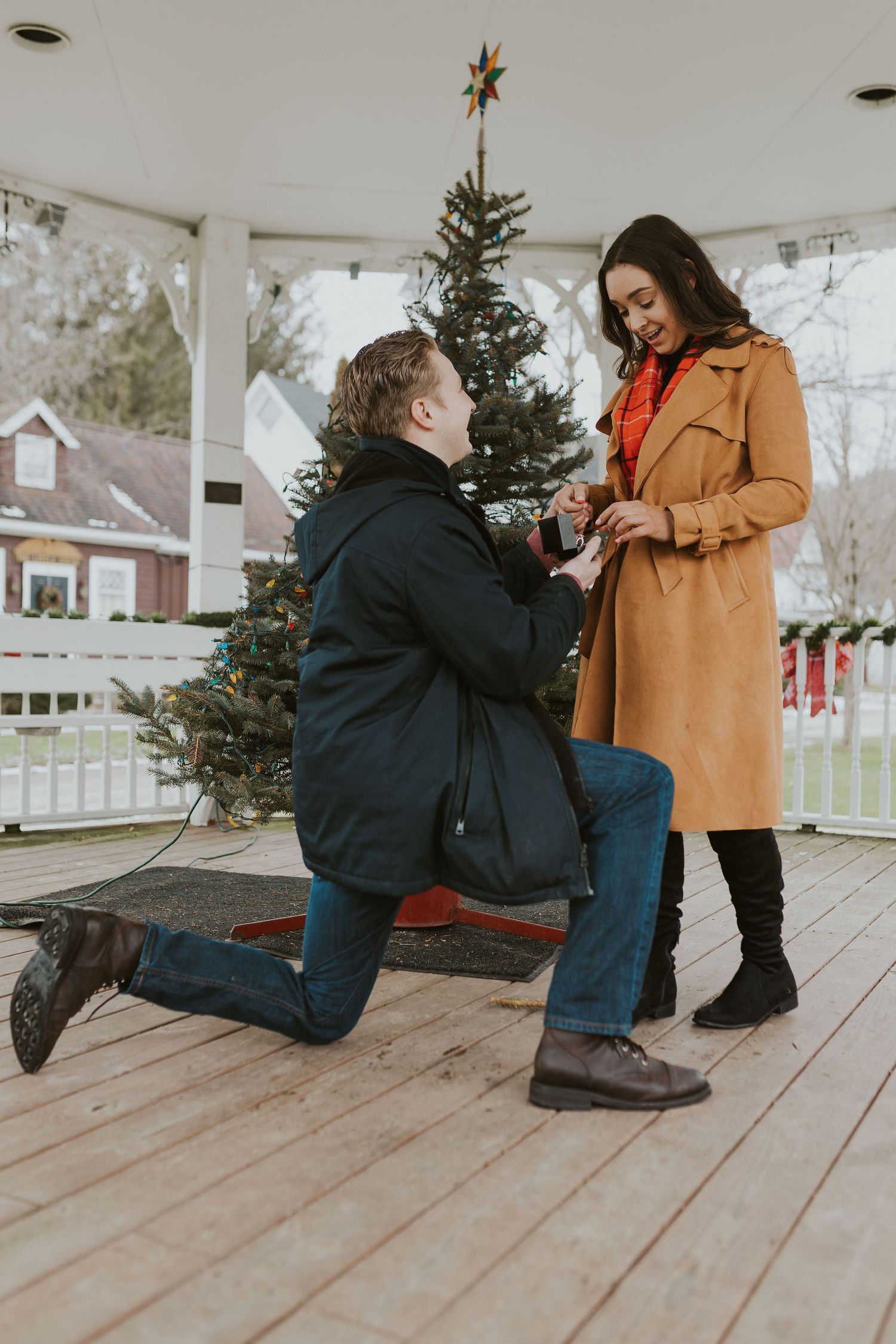 Man proposing on one knee to woman in gazebo with Christmas tree backdrop.